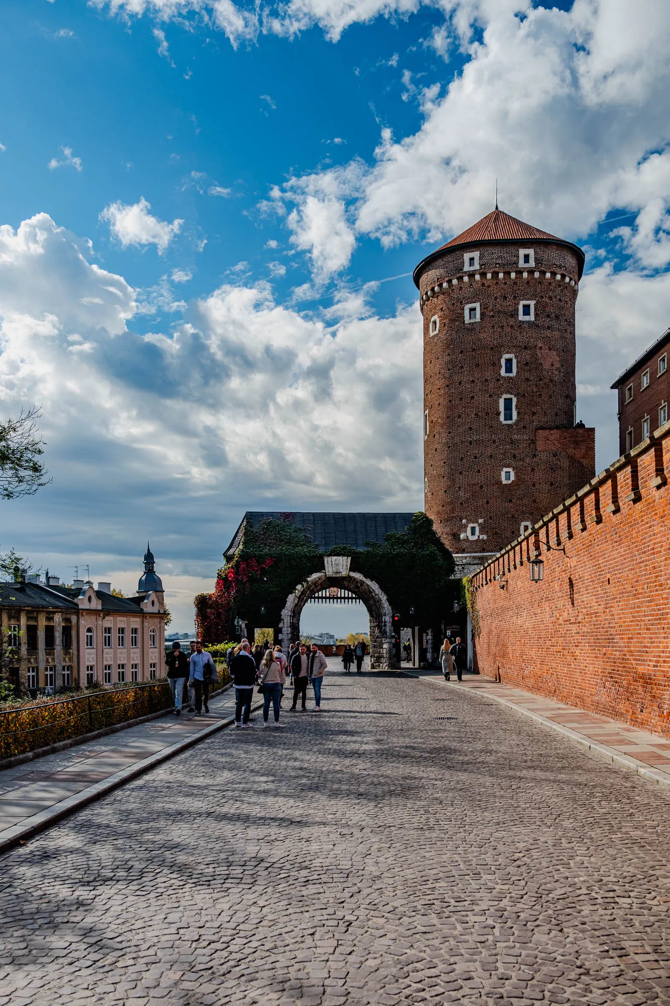 The image shows a cobblestone path leading towards a large, brick archway. The archway is covered in green vines and has a large iron gate. To the right of the archway is a tall, red brick tower with a conical roof. The tower is part of a larger brick wall that extends out of the frame. In the distance, there is a smaller building with a pointed roof. There are a few people walking on the path, and the sky is blue with fluffy white clouds.