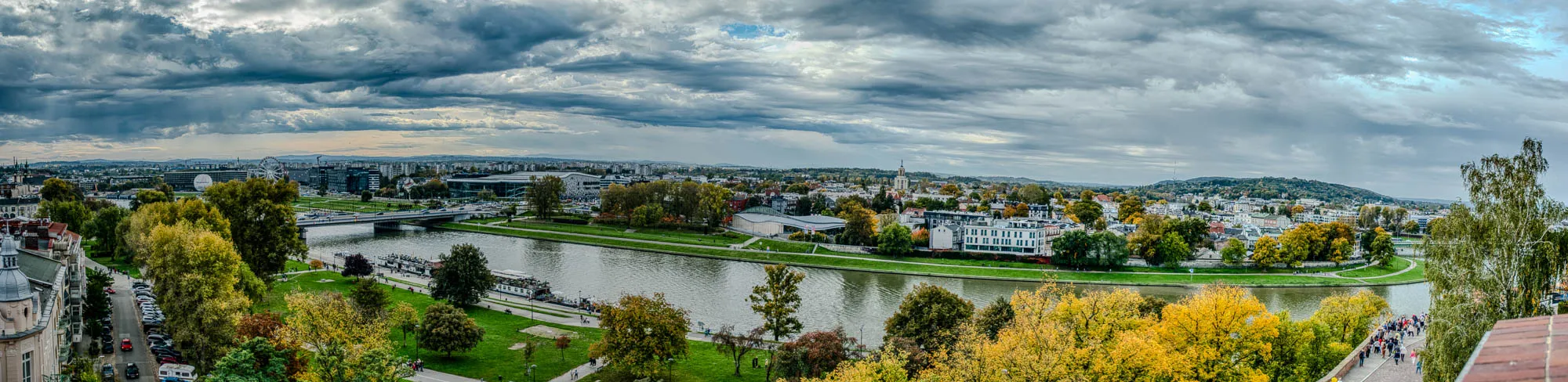 This is a panoramic image of a city with a river running through it. It’s a sunny day with some clouds in the sky. The river is winding and the cityscape is in the distance. There are green trees and grassy hills lining the river. You can see a bridge over the river to the left. There’s a large white building in the center of the image with a tall steeple on top. To the right, a group of people is walking along a pathway that leads to the top of the hill. There’s a red brick wall on the lower right of the image. 
