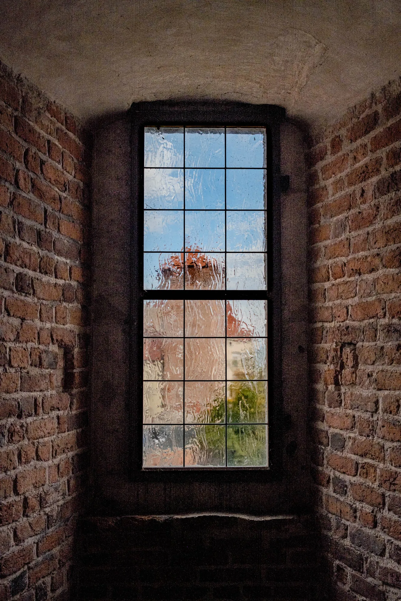 A window with a dark brown frame is set into a brick wall. The window is made up of nine panes of glass with black lines between them. Through the wavy glass you can see a blurred view of a building with a red roof and green trees beyond. The brick wall is made up of dark red and brown bricks. The wall above and below the window is mostly dark with hints of a light brown color. The background is dark and unidentifiable.  