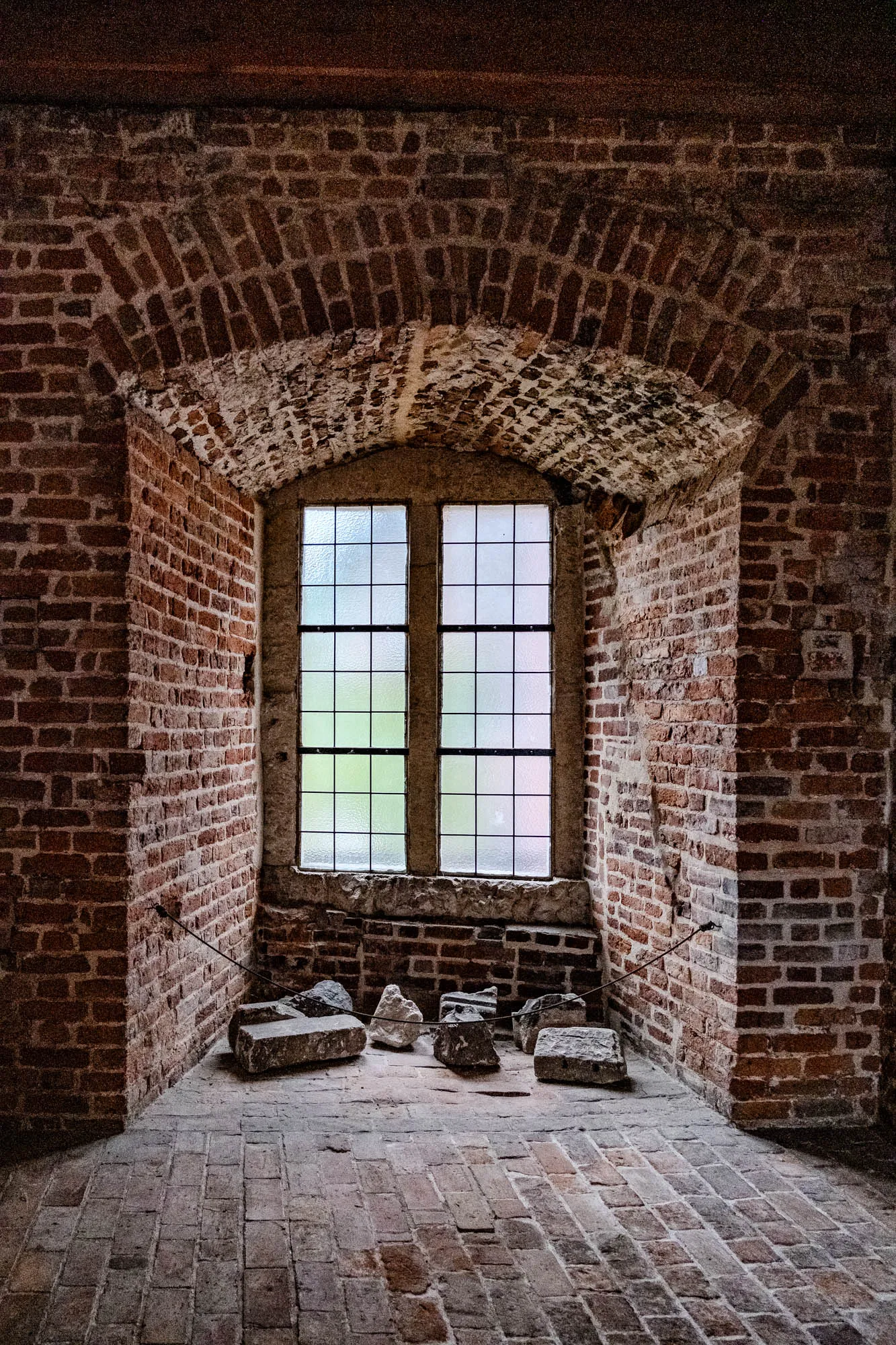 The image shows a brick room with a large window. The brick wall is made of red bricks laid in a pattern. The bricks are worn, creating a rough texture. The window has a large pane of glass divided by a grid of thin black lines into 12 squares. The window is slightly arched at the top, like a doorway. The room is dimly lit, and the window is the main source of light.  There are four large rocks scattered on the floor in front of the window. The floor is made of brick, arranged in a pattern of long, thin rectangles.