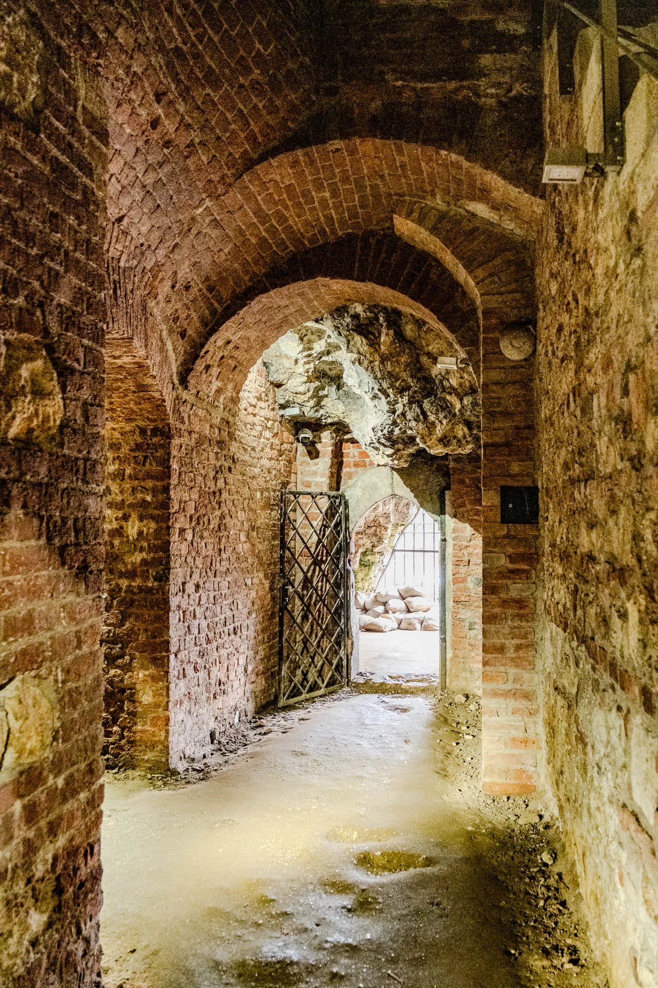 The image shows a narrow brick hallway with an arched ceiling. The hallway is lit by a single window at the end, which is a doorway leading to another area. The doorway is framed by two iron gates. There are a few large sandbags visible in the doorway. The floor of the hallway is dirt and uneven. The walls are made of brick and are rough and textured. The ceiling is made of stone and is also uneven. The hallway is dark and shadowy.