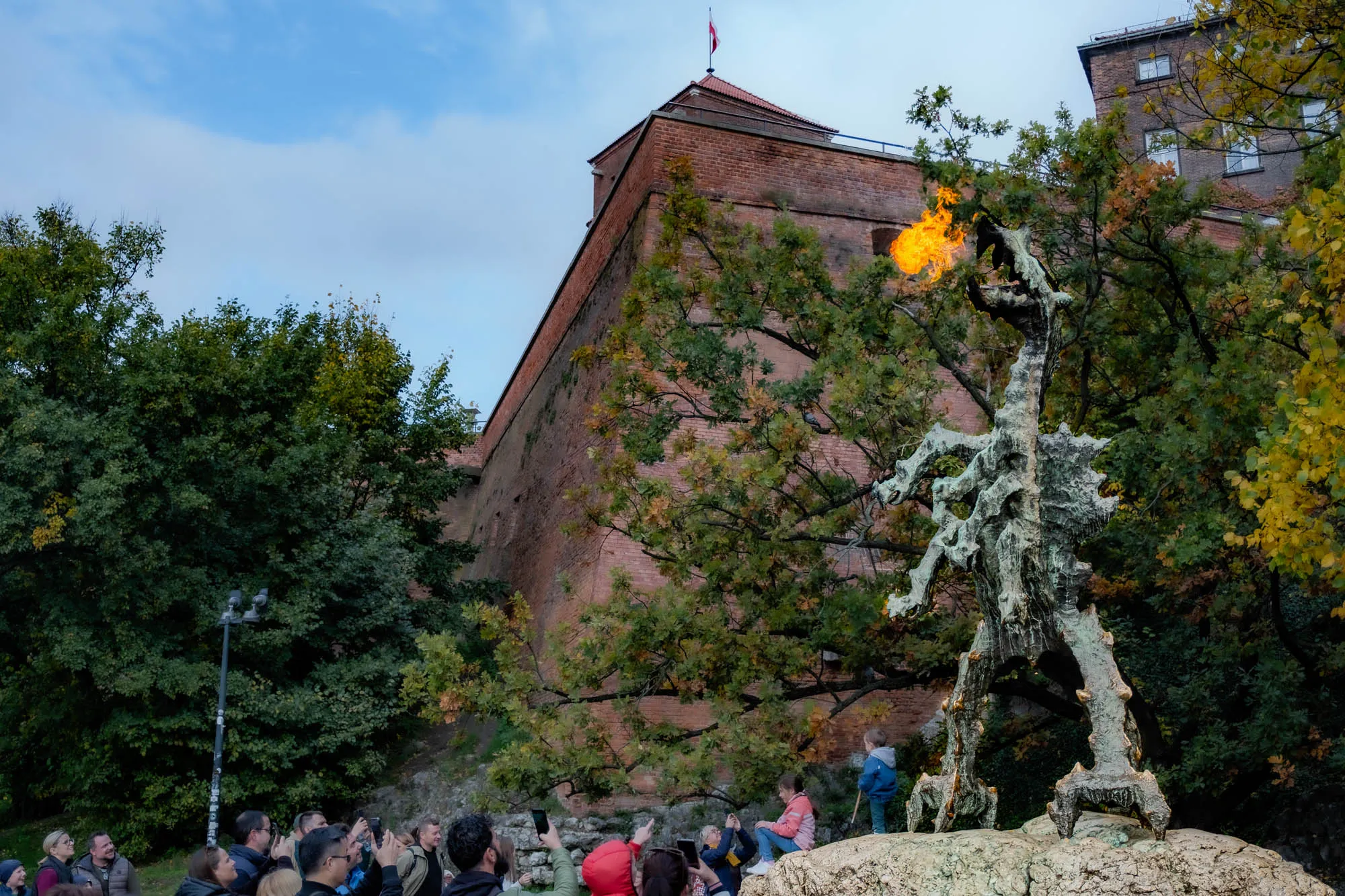 The image shows a large, bronze statue of a dragon. The dragon is situated on a rocky outcrop in front of a brick building, and a tree is to the right of the dragon, obscuring the building. The dragon is in a rearing posture, with its head raised and its mouth open. It appears to be breathing fire as the opening of its mouth is lit with a bright orange flame. 

The dragon is in the foreground of the image, while the brick building is in the background. The brick building is tall and has a flat roof. A flagpole is visible on top of the building, and a flag hangs from it. The flag has a white and red design on it. 

The dragon is surrounded by trees and shrubs, and a group of people are standing in the foreground, looking up at the dragon and taking pictures. 

The sky is blue and partly cloudy. 
