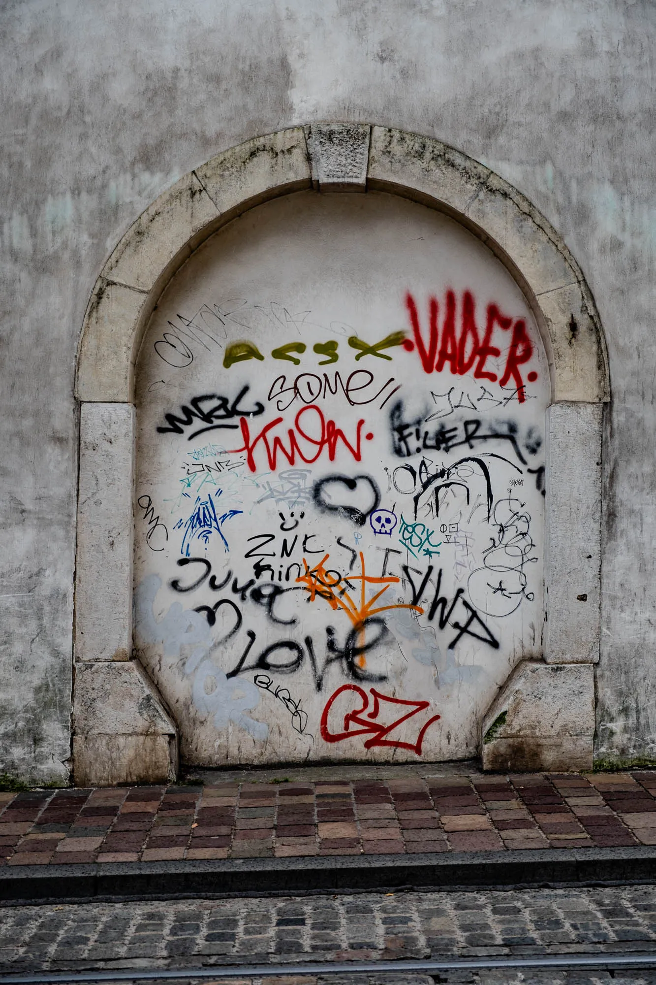 The image shows a brick archway in an old, grey stone wall covered in graffiti. The brick archway is painted white and has a variety of graffiti on it, including the word "Vader" in red, the words "WAB SOME" and "KNOW" in black and red, the word "LOVE" in black, and the word "GZ" in red. There are also various symbols and squiggles in black, blue, orange, yellow, and green paint. The image is taken from a low angle, and the brick archway is in focus while the wall is out of focus. In front of the archway is a brick walkway that leads to a cobblestone street. There is a metal track embedded in the cobblestone street.  There is moss growing on the base of the wall.