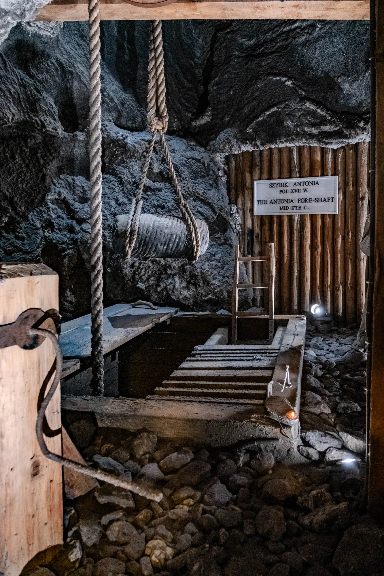 This is a photo of a mine shaft in a salt mine. It appears to be an old mine as the walls are made of rough-hewn wood and the shaft itself is fairly narrow. A thick rope is hanging from a wooden beam across the top of the shaft. The rope is attached to a large, round, heavy object. The bottom of the shaft is covered with loose stones.  A small wooden ladder leans against the wall next to a sign that reads, "Szybik Antonia Pol XVII W. The Antonia Fore-Shaft Mid 17th C." The sign appears to be white with black lettering on it.  There is a soft light shining from the bottom of the shaft and a small light on the wall.