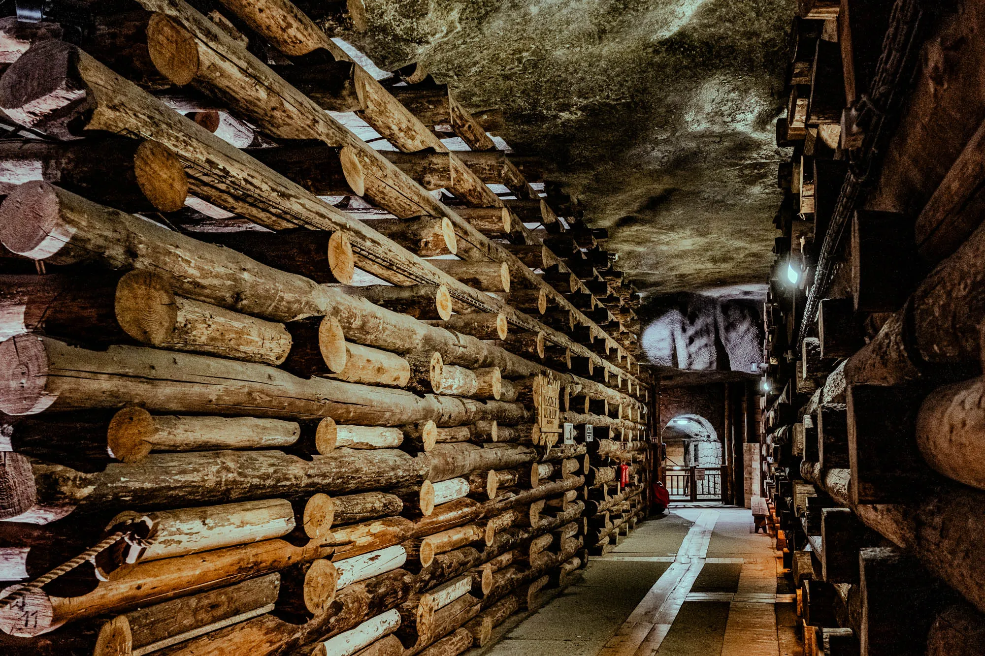 The image shows a tunnel lined with logs stacked from floor to ceiling. The logs are mostly brown and some have a white or gray patina. The logs are stacked in a staggered pattern and the ends of the logs can be seen in the image. The tunnel appears to be dimly lit, with light coming from the end of the tunnel. At the end of the tunnel is a wooden walkway with a metal railing. The wall behind the walkway appears to be made of stone. The tunnel may be an old mine shaft or a timber-framed structure. The logs are arranged in a way that gives the impression of an open-air tunnel. The logs are old and worn, adding to the feel of the tunnel.