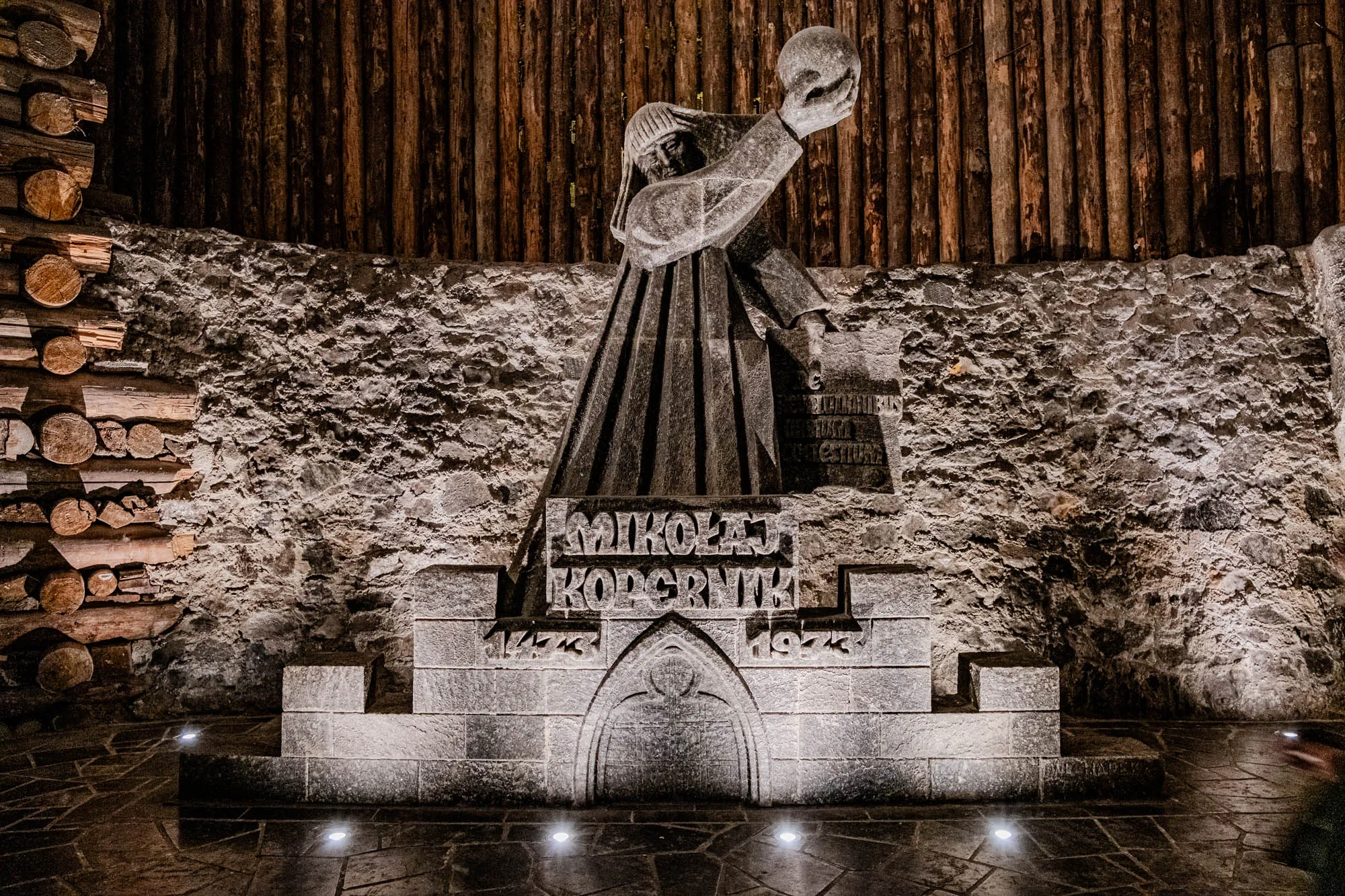 This is a photo of a stone statue of a man holding a globe. It is set into a stone wall with stacked logs to the left of the sculpture. The statue is wearing a long robe and has a serious expression. The base of the statue has the words "MIKOŁAJ KOPERNIK" in block letters, along with the years "1473" and "1543." There are lights illuminating the area and the statue.