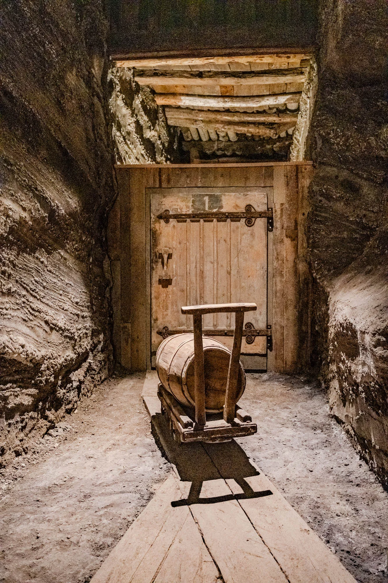 The image shows a narrow, dimly lit corridor with rough, textured walls. At the end of the corridor, there is a wooden door with heavy metal hinges. The door appears to be closed. In front of the door, there is a wooden barrel mounted on a wooden frame. The barrel is facing the viewer and sits on a wooden platform. There are two wooden beams overhead, providing the only light source.  The corridor floor is dirt and looks uneven. The image is likely taken from a low angle, as the barrel dominates the foreground.  The corridor is likely in an old mine, or an underground space with a rustic, historical appearance.  The image creates a sense of mystery and anticipation, leaving the viewer wondering what lies behind the door.