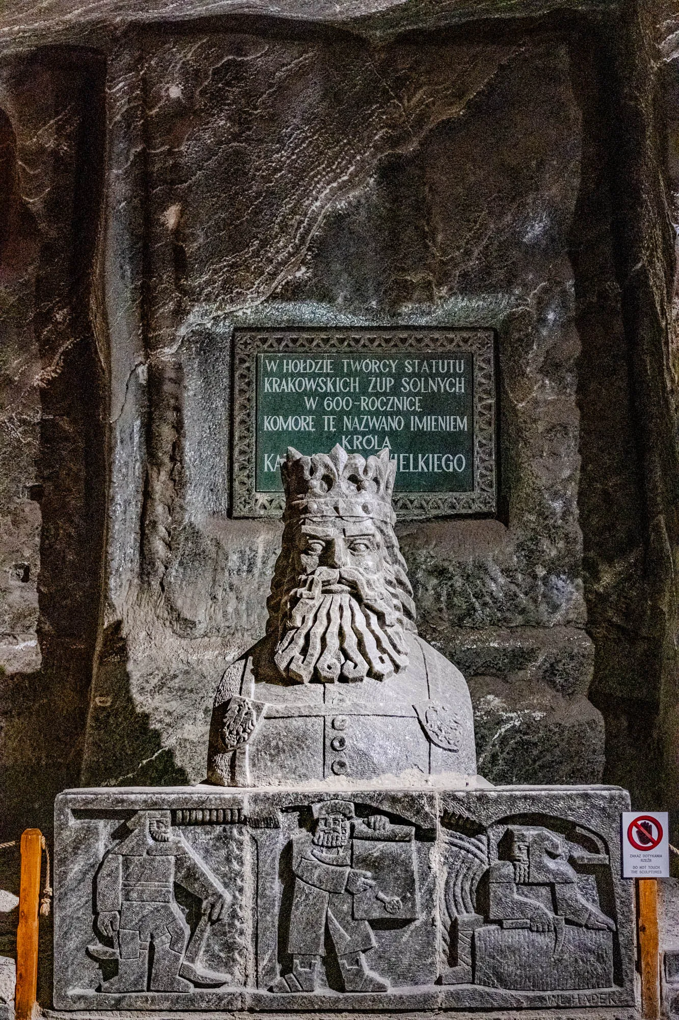 The image shows a carved bust of a man with a crown on his head and a long beard. He is sitting on a carved stone throne. The throne has carvings of three men in work clothes holding pickaxes. Above the bust is a rectangular stone plaque with black lettering.  The lettering is in Polish and reads: "In honor of the creator of the statutes of the Krakow Salt Works, on the occasion of the 600th anniversary, this chamber was named after King ..." The last part of the inscription is illegible. The plaque is mounted on a rough stone wall.  To the right of the image is a red and white sign that reads: "No touching carvings" in both Polish and English.