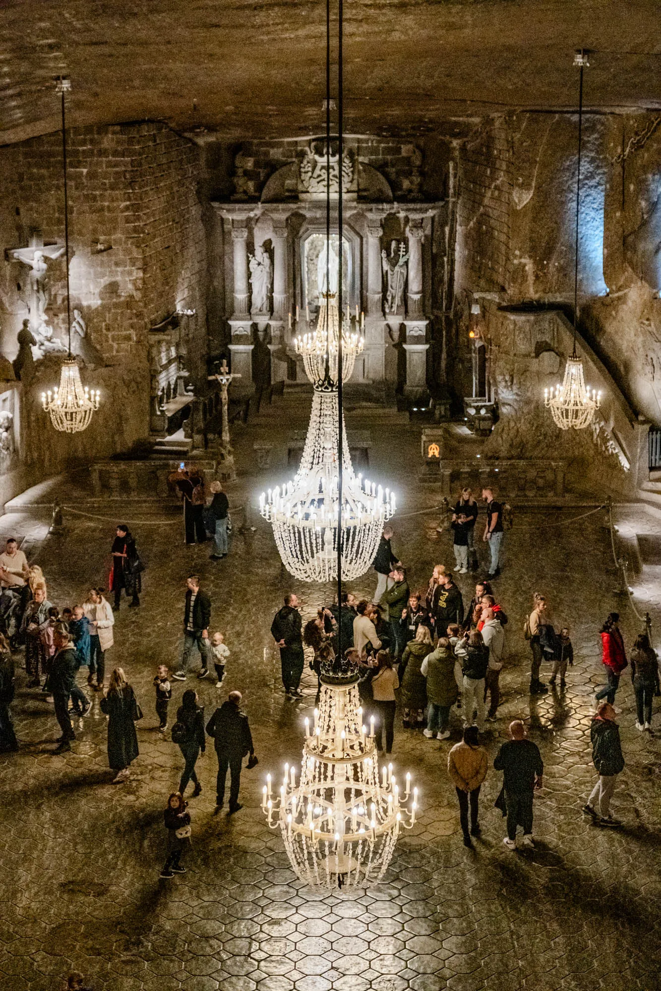 The image shows a large, open underground space with stone walls and a cobblestone floor.  In the center of the image hangs a large, ornate chandelier, surrounded by smaller chandeliers, with a group of people standing around them.  The space appears to be a chapel, with an altar in the back, a crucifix hanging on the wall, and statues on either side of the altar. The walls are rough-hewn stone, and the space is dimly lit by the chandeliers. The people are standing in groups, and some are looking at the chandeliers or taking pictures.  There is a sense of awe and wonder in the air, as people admire the beauty of the space.