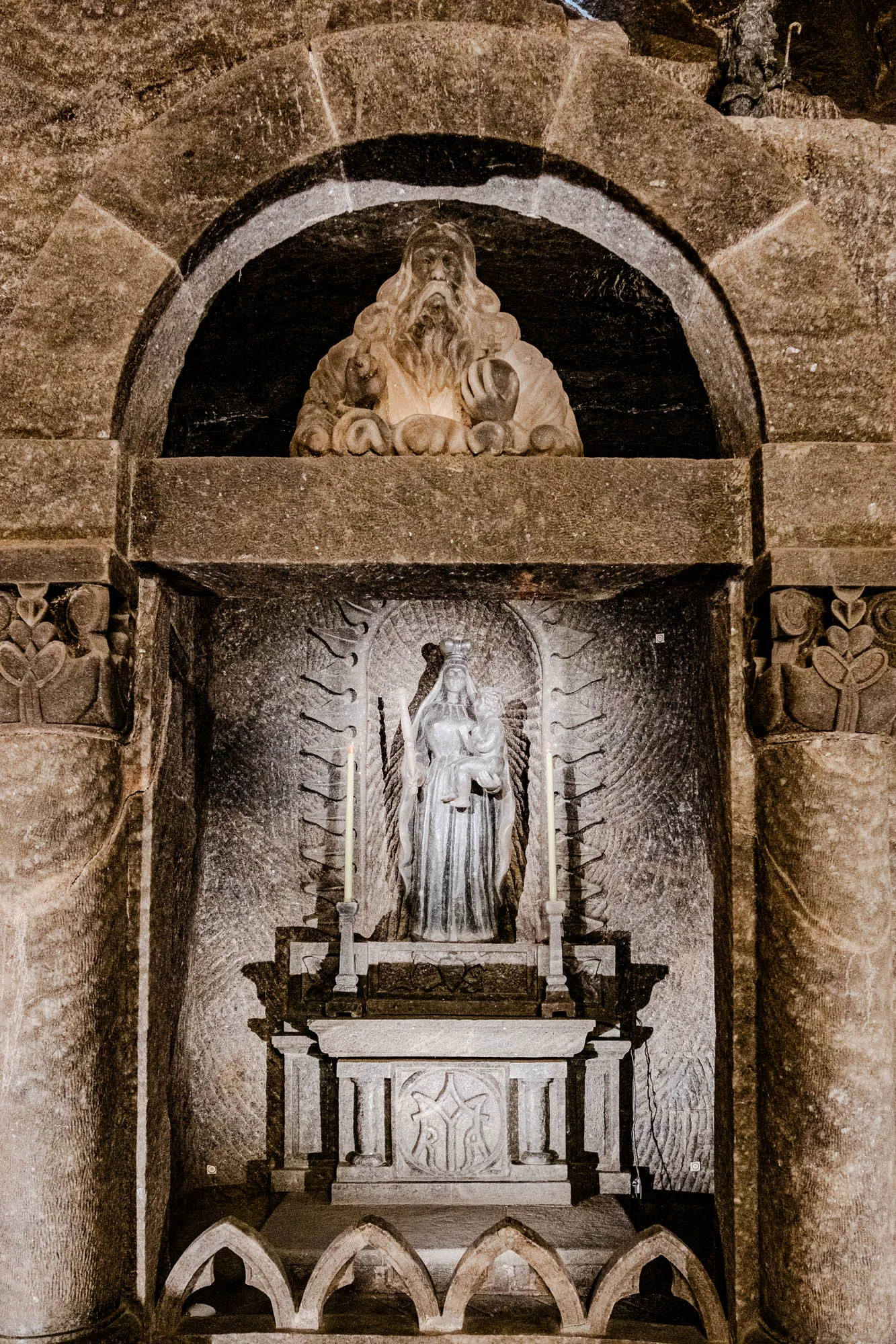 This is a photograph of a shrine in a dark cave. The shrine is made out of stone. There is a figure of a woman holding a child in her arms on top of a stone altar. The woman is standing on a pedestal and has a crown on her head. There is a candle on each side of the woman and a carving of an angel with outstretched wings behind her. At the top of the shrine, there is an archway with a carving of a man with a long beard and hair. The background of the photo is very dark and is made up of rough stone.  This is a very dark and mysterious picture.  The stone is rough and looks like it is made of salt.