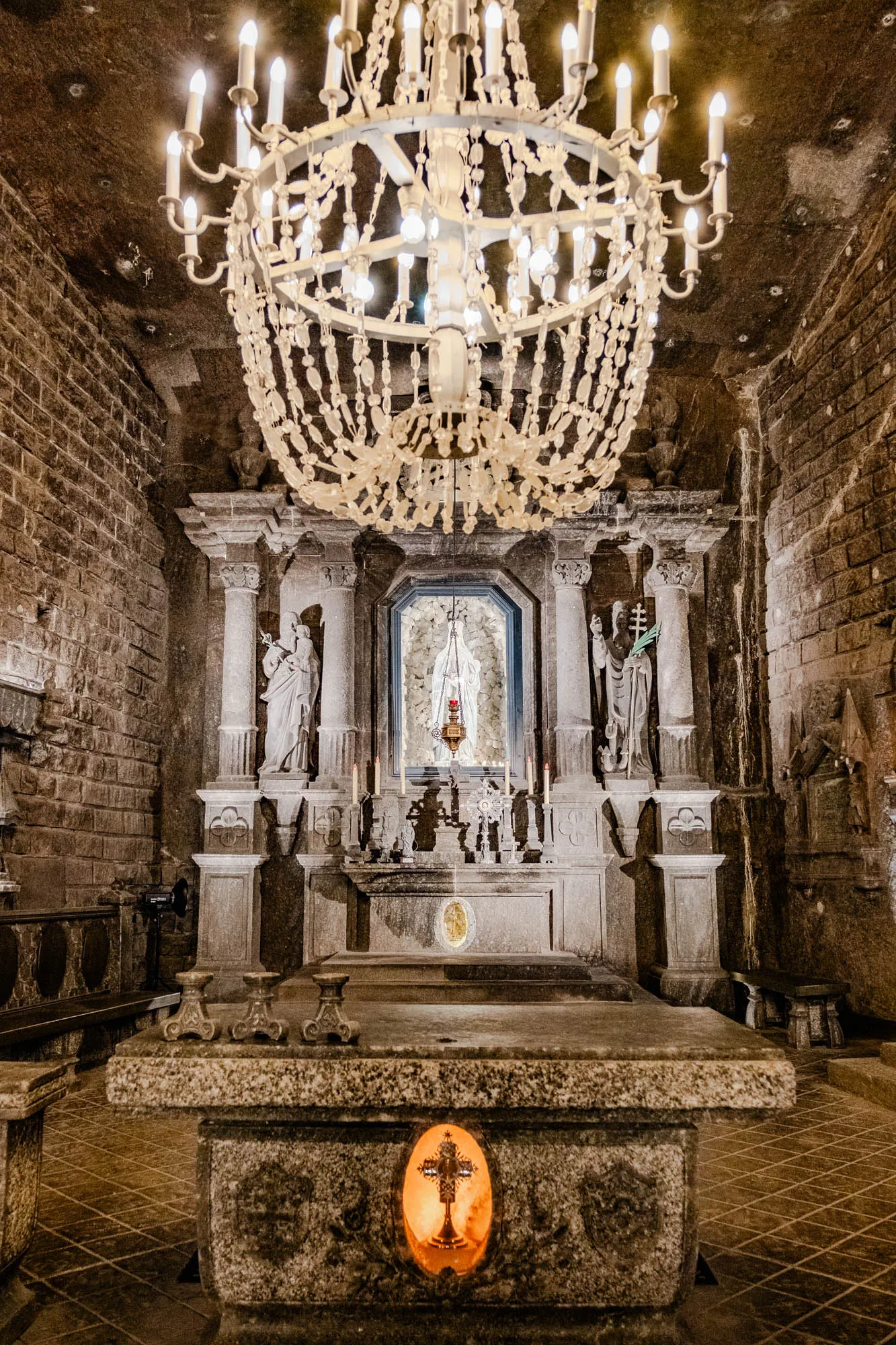 The image shows a small, stone chapel carved out of a salt mine. A large, white crystal chandelier hangs from the ceiling. The chapel is dimly lit, and the walls are rough and uneven. There is a stone altar with a statue of the Virgin Mary in a recess behind it. The altar is flanked by two stone columns topped with sculptures of two figures. In front of the altar, there is a stone table with a recessed opening in the center. A golden cross glows brightly within the opening. The walls of the chapel are lined with benches.