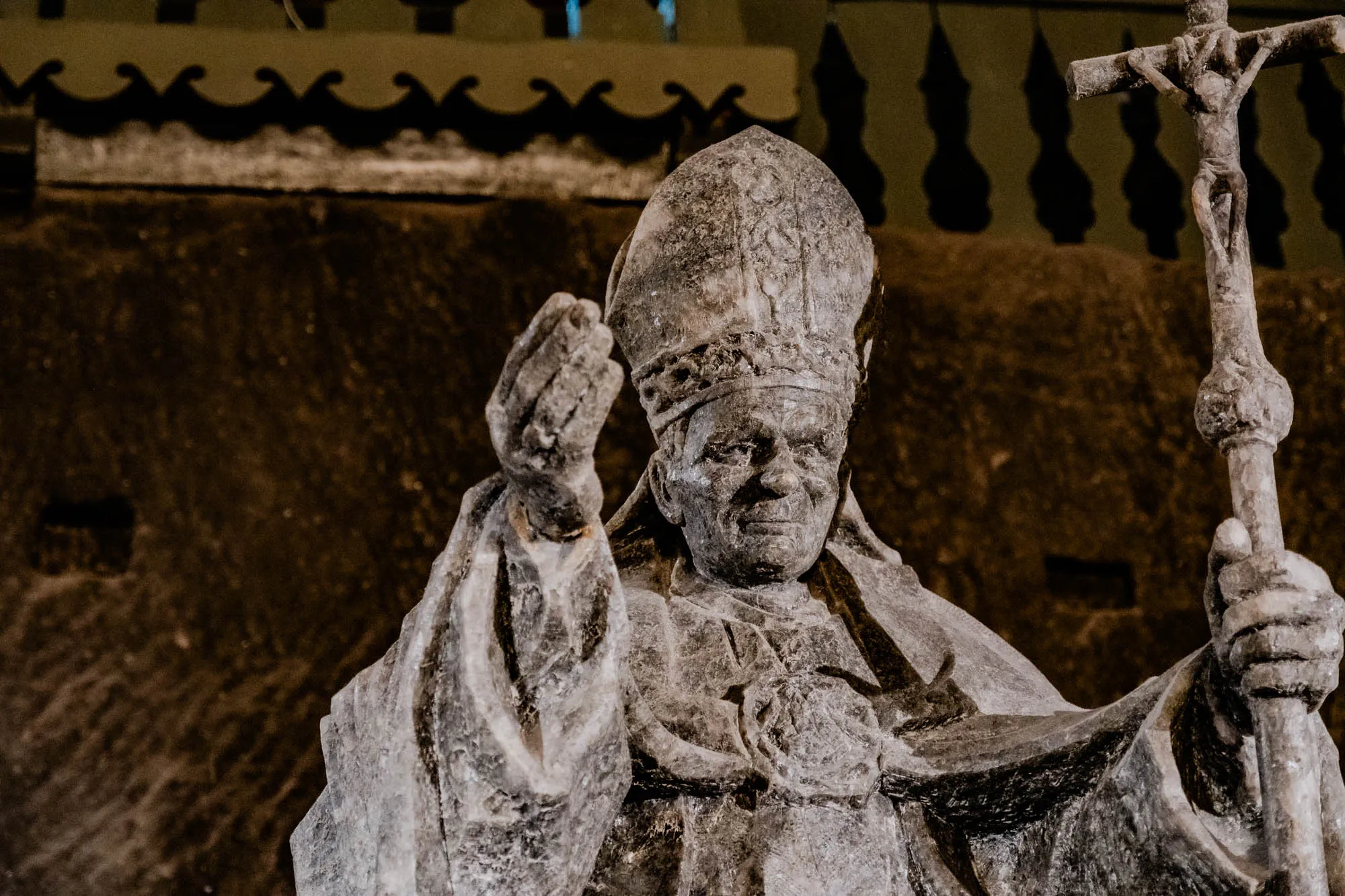 The image shows a salt statue of a man wearing a hat and robes, standing in front of a salt sculpture of a cross. The man's right hand is raised, and he is holding a staff in his left hand. There is a dark, rough, textured wall behind the sculptures, suggesting a salt mine.  The image has a dim lighting, possibly coming from above the statues. The sculptures are detailed and look realistic, with a textured surface that is likely made of salt. The cross sculpture has a figure of Jesus Christ on it.