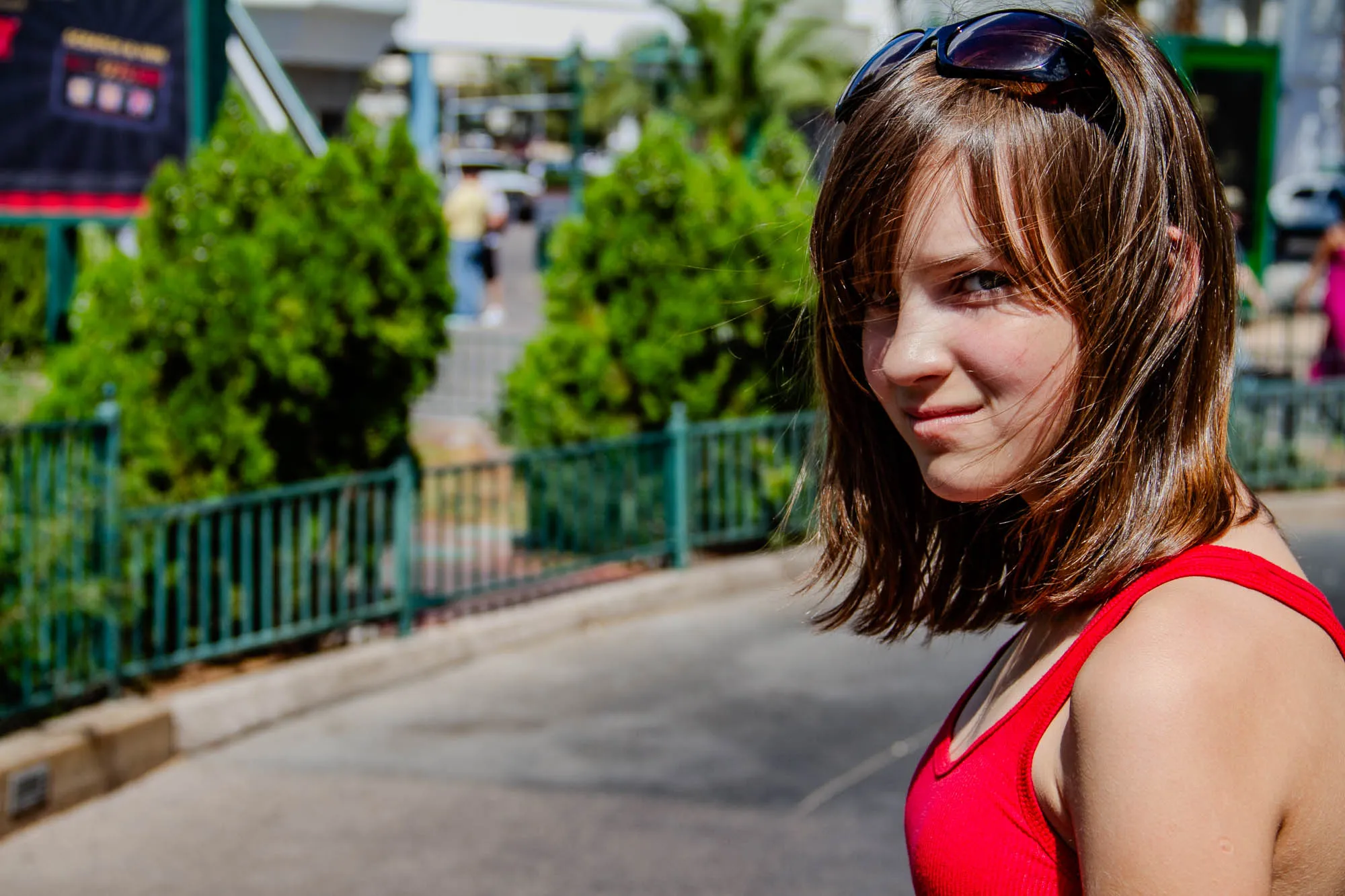 The image shows a young woman with long, brown hair, standing in front of a green fence and a paved road. The woman is wearing a red tank top and black sunglasses on top of her head, which are pushed back on her forehead. The woman is looking directly at the camera and smiling slightly. The background is blurry, suggesting that the image was taken with a shallow depth of field.  There are green bushes behind the fence.  