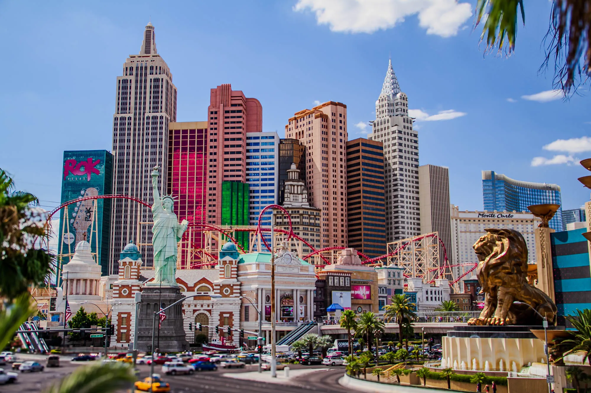 A wide-angle view of the New York, New York hotel in Las Vegas, showing a condensed representative skyline of Manhattan, new York. The Chrysler Building and the Empire State Building are specifically represented, as is the Statue of Liberty standing in front. A red roller coaster runs behind the Statue of Liberty and the buildings. In the foreground to the right is a giant golden MGM lion, surveying the street both hotels share.