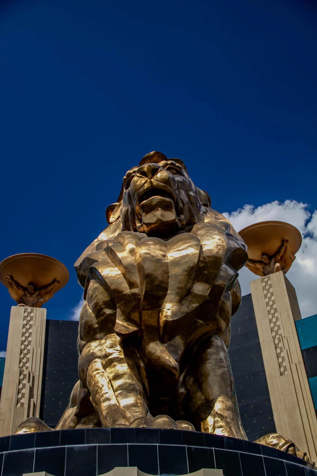 A large golden lion statue is sitting on its haunches with its mouth open in a roar. It's facing the camera and looking upward at the sky. The statue is in front of a building, likely a casino, with a large, flat, golden roof that is being held up by a man in a similar pose to Atlas, with his back to the viewer. The sky is a bright blue with a small, fluffy white cloud to the right of the lion.  The lion's face is detailed and looks somewhat angry.  The lion's body is sculpted in a manner that makes it look like its fur is flowing, particularly around its chest. There is a black tiled area in the foreground.