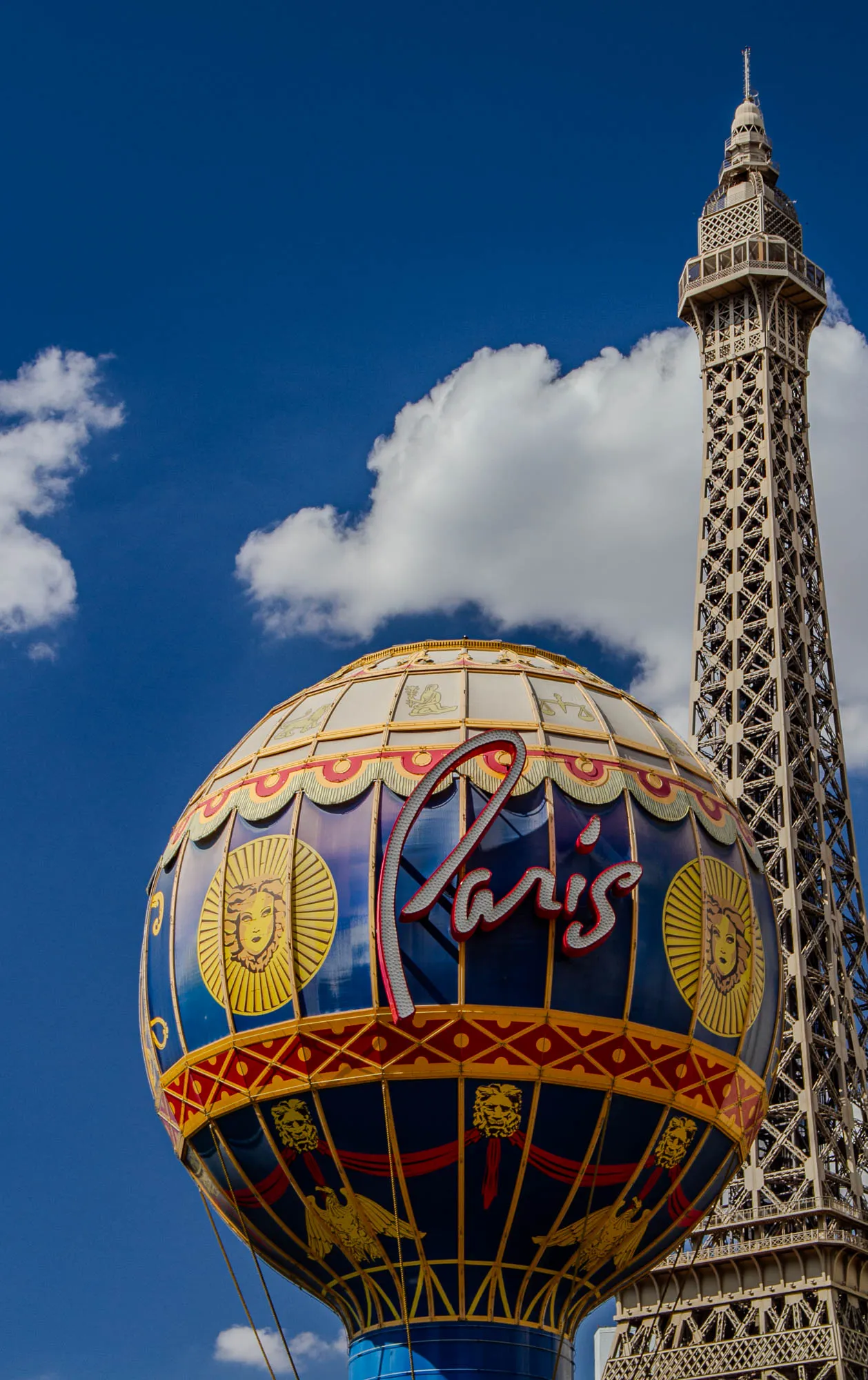 The image shows a hot air balloon in the shape of a globe, with the word "Paris" written in red, white and blue letters on it. The balloon has blue, yellow, and red stripes with images of lions and eagles surrounding it. It is positioned in front of a brown metal Eiffel Tower replica. There is a blue sky with white clouds in the background.