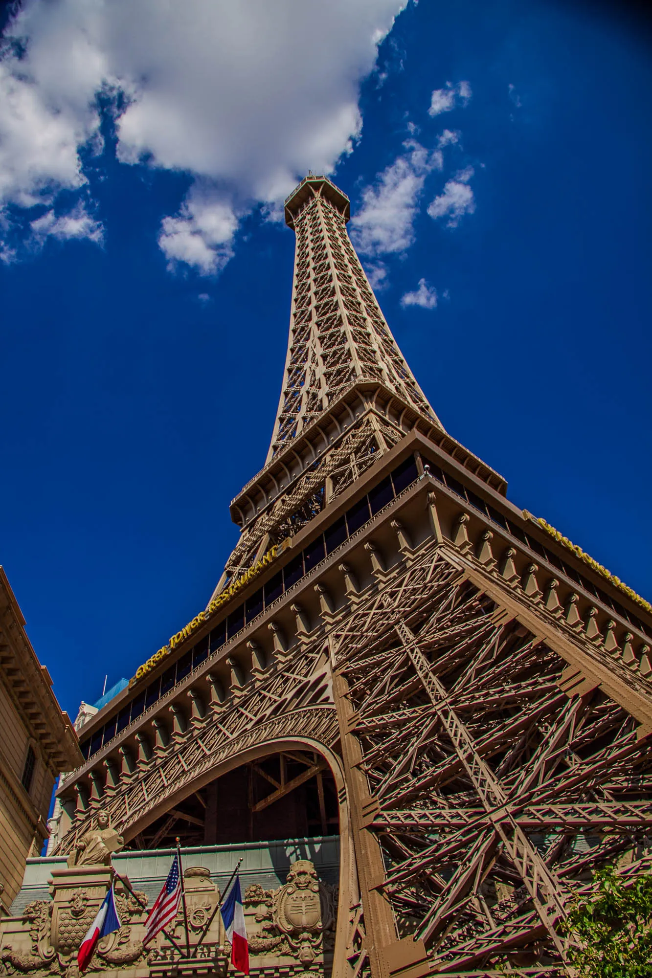 The image is a low-angle shot of the Eiffel Tower replica in Las Vegas, Nevada. The tower is made of metal and rises up towards the bright blue sky. There are white clouds in the sky, and the top of the tower is out of frame.  You can see the intricate metal framework of the tower, and there are some windows and balconies visible. At the bottom of the tower, there are two flags hanging from the structure: a French flag and an American flag. The tower's name "Eiffel Tower Restaurant" is partially visible in yellow lettering.  The image is taken from a ground-level perspective looking up at the tower. The base of the tower is partially obscured by some green bushes.