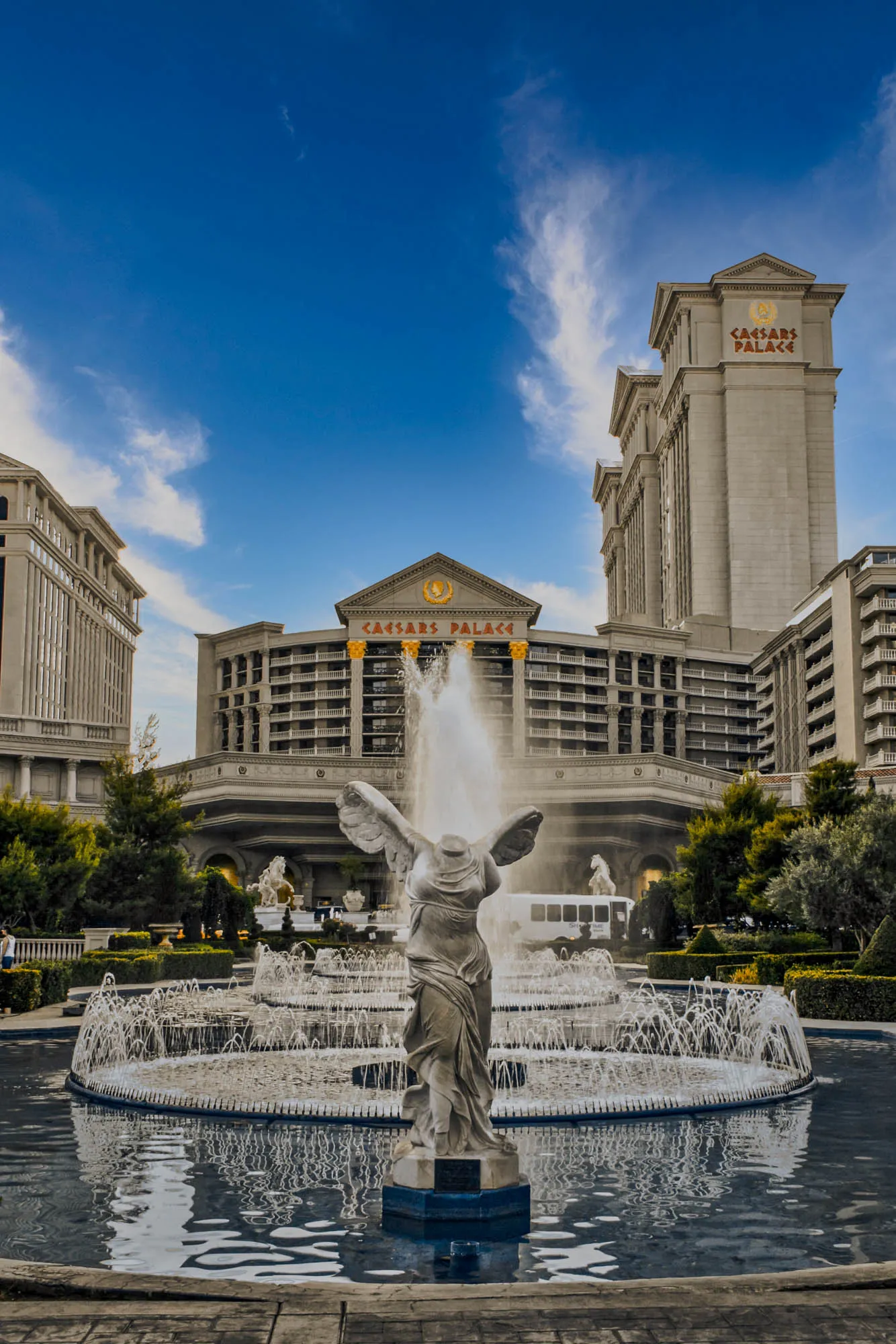 The image depicts a large fountain in front of Caesars Palace in Las Vegas. The fountain has multiple tiers and is shaped like a circle. It has a statue of a woman with wings standing in the center. There are several water jets shooting water into the air. In the background, there are two tall buildings with the words "Caesars Palace" written on the top.  The sky is blue with white fluffy clouds.  The image is taken from a low angle.