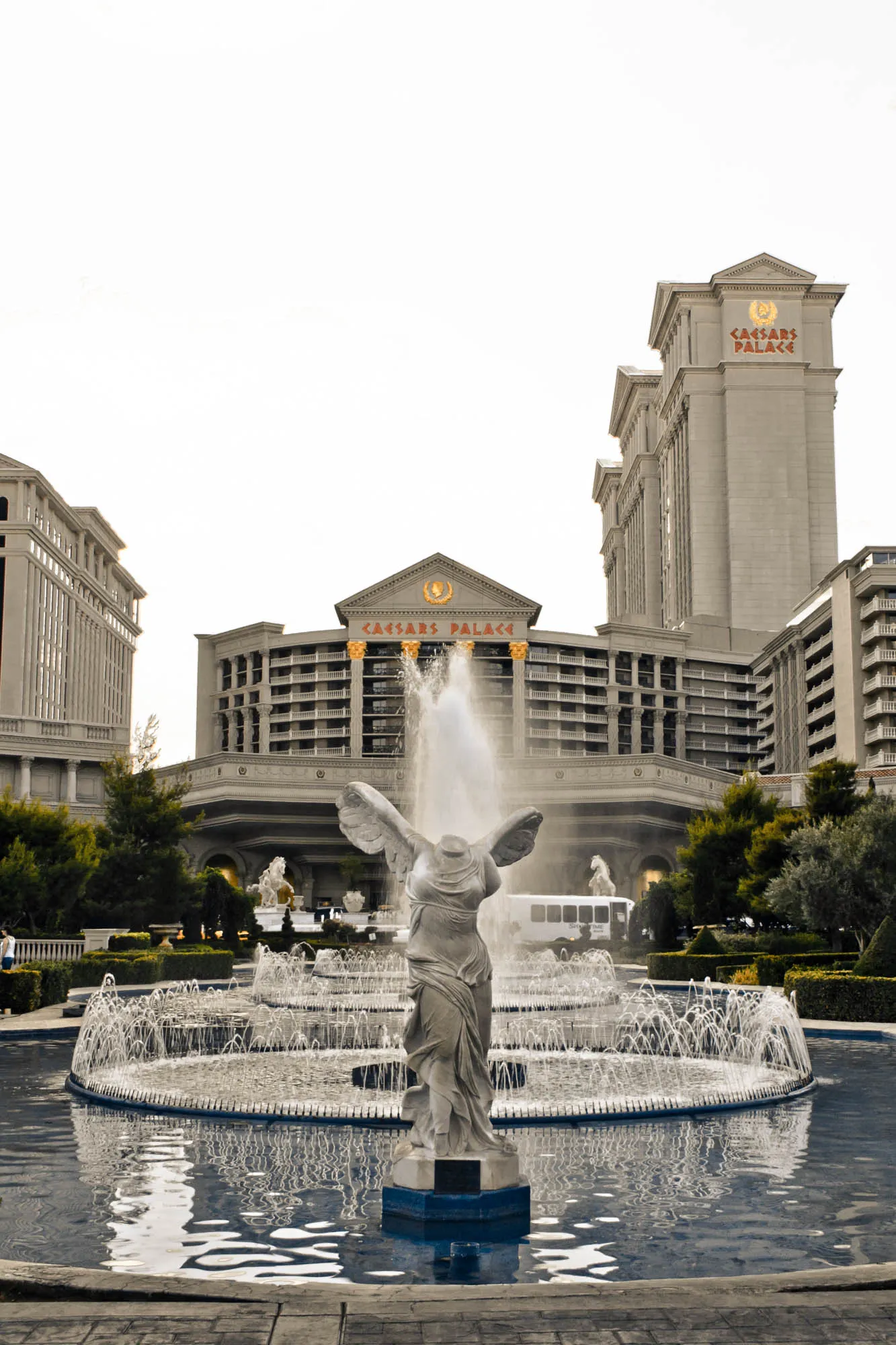 The image is of a large fountain in front of Caesar's Palace in Las Vegas. The fountain is circular and has many jets of water spraying up in the air. In the center of the fountain is a statue of a woman with her arms outstretched, it's difficult to tell what she is holding as her arms are broken off at the elbows.  The statue is on a pedestal surrounded by the fountain. Behind the fountain is a large, modern building with a sign that reads "Caesar's Palace". The building appears to be several stories high and is made of beige stone. There are trees and bushes in front of the building, and there is a clear blue sky above. There are a couple of other buildings visible in the background as well.