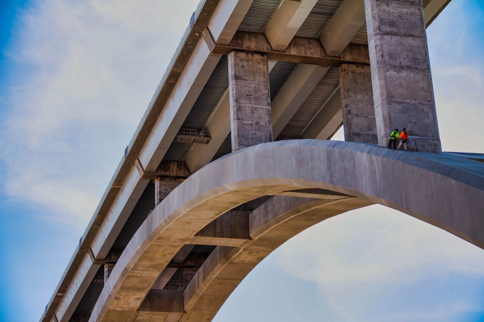 The image shows a concrete bridge with two workers standing on the upper deck. The bridge is made up of a flat, horizontal deck with a curved arch underneath. The underside of the flat deck is visible, with multiple concrete pillars and a light gray, corrugated surface. The workers are standing on the flat deck, facing the viewer, with the arch and underside of the deck behind them. The sky is a light blue with white clouds, visible through the open space of the arch.  The workers are wearing safety harnesses and appear to be working on the bridge.