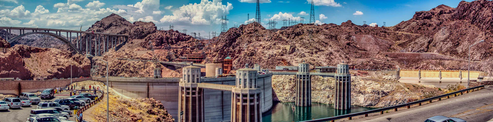 A wide panoramic view of a dam on a sunny day. The dam is made of concrete and has a large arched bridge over the top of it. The bridge is supported by a series of concrete pillars. The dam is situated in a rocky canyon. There are several large power lines that run along the sides of the canyon. There are several cars parked in a parking lot in front of the dam, along with a few people standing around.  The dam is partially submerged in a body of water. The sky is blue with white clouds.  There are several large rock formations in the background.  There are also some buildings in the background, including a building with a red roof.