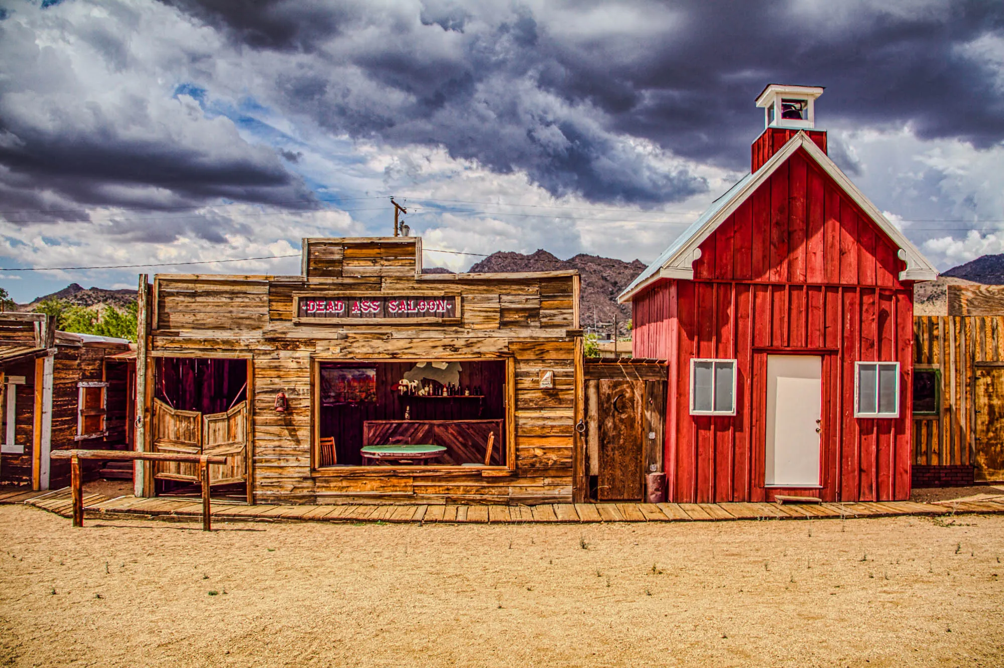 This image depicts a scene from the Wild West. In the foreground is a sandy expanse, bordered by wooden walkways. There are three buildings in the image: a saloon with the words "DEAD ASS SALOON" in red above its entrance; a small, red building with a white door and two small windows; and a third, larger, wooden building partially obscured by the red building. The saloon is made of wood planks and has a saloon door at the entrance. The window is open, and the interior is visible. The red building is simple and has a bell tower on top. The wooden building is also made of wood planks and has a small window. Above the buildings, there is a cloudy sky with a hint of blue peeking through.  The scene is set against a backdrop of a mountain range, partially obscured by the buildings.  