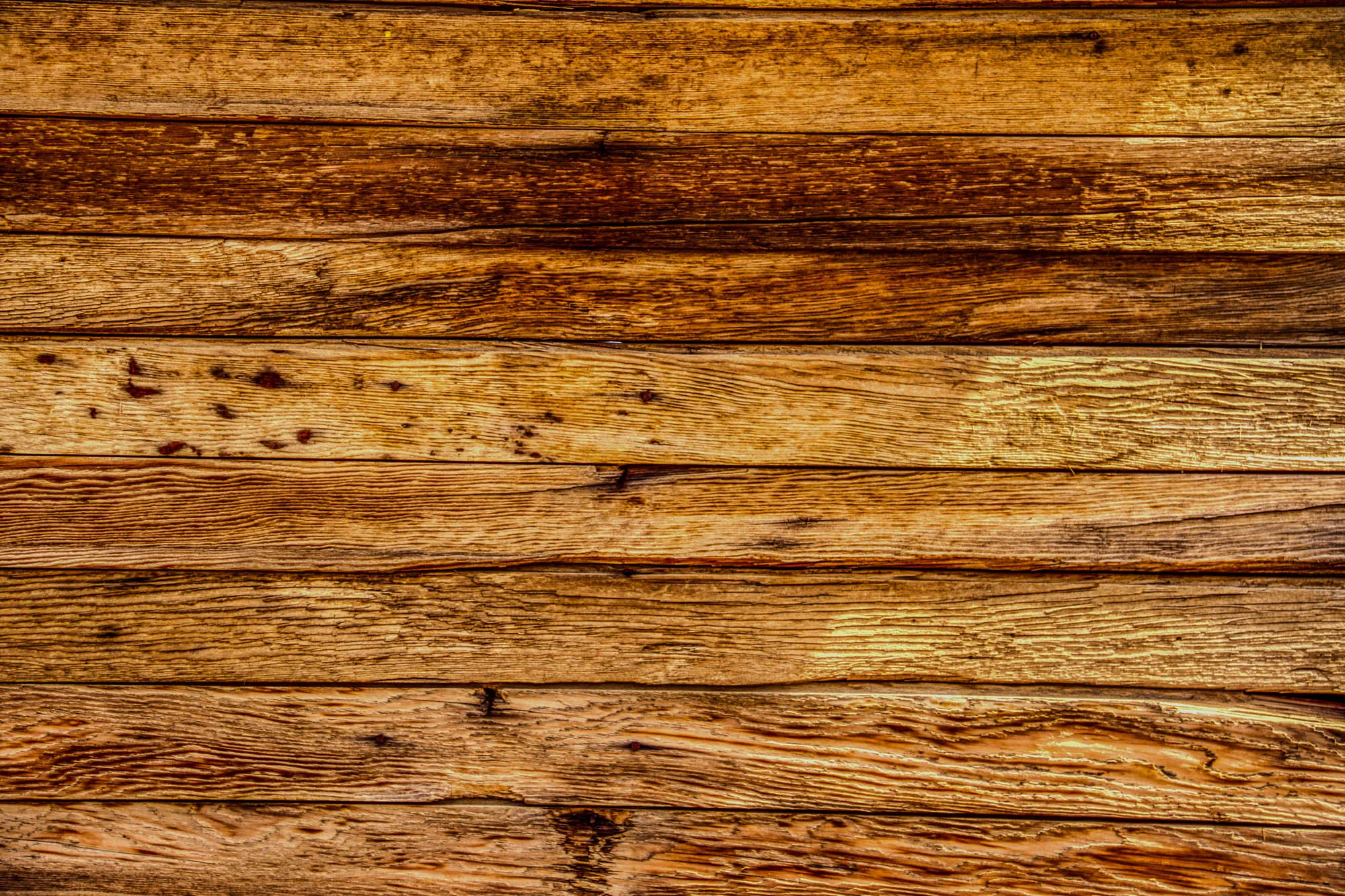 The image is a close-up of a wall made of horizontal wooden planks. The wood is old and weathered, with a lot of grain showing. The planks are of different widths and have a range of shades of brown, from light to dark. There are some knots and imperfections in the wood, and a few nail holes. The image is likely intended to convey a sense of rustic charm and natural beauty.