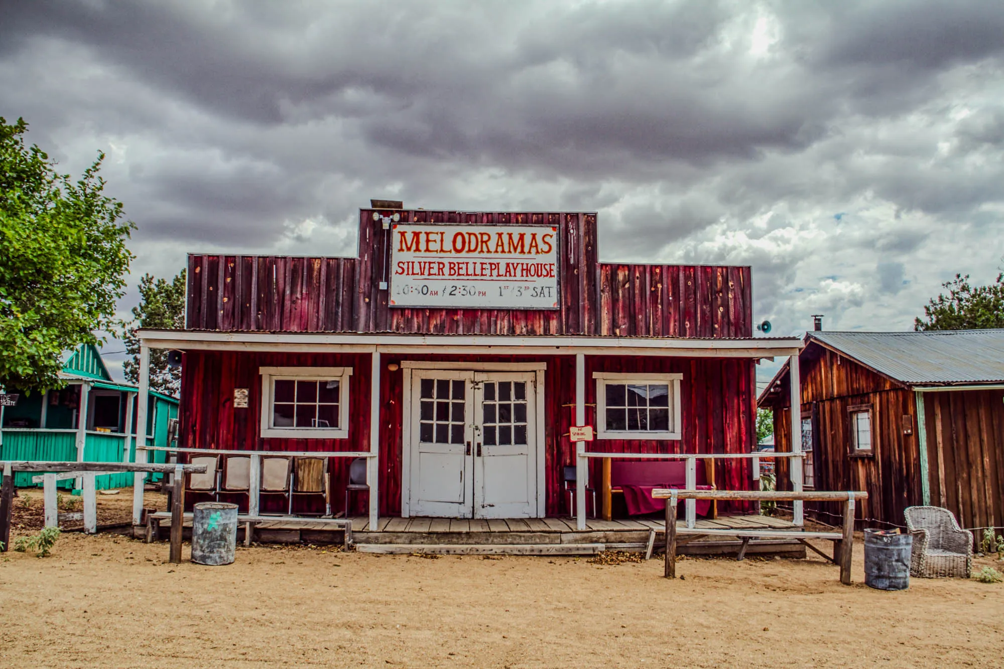 A red wooden building with a white trim and a sign above the entrance that reads "Melodramas Silver Belle Playhouse 1:30am 2:30pm 1:30 Sat". The building has a wooden porch with white railings and a wooden bench on the right. The building is situated in a dusty area with a cloudy sky above. A second, smaller, brown building with a metal roof is to the right of the main building. A  metal trash can is in front of the main building, a wooden bench is next to the trash can, and a wicker chair is in front of the smaller building.