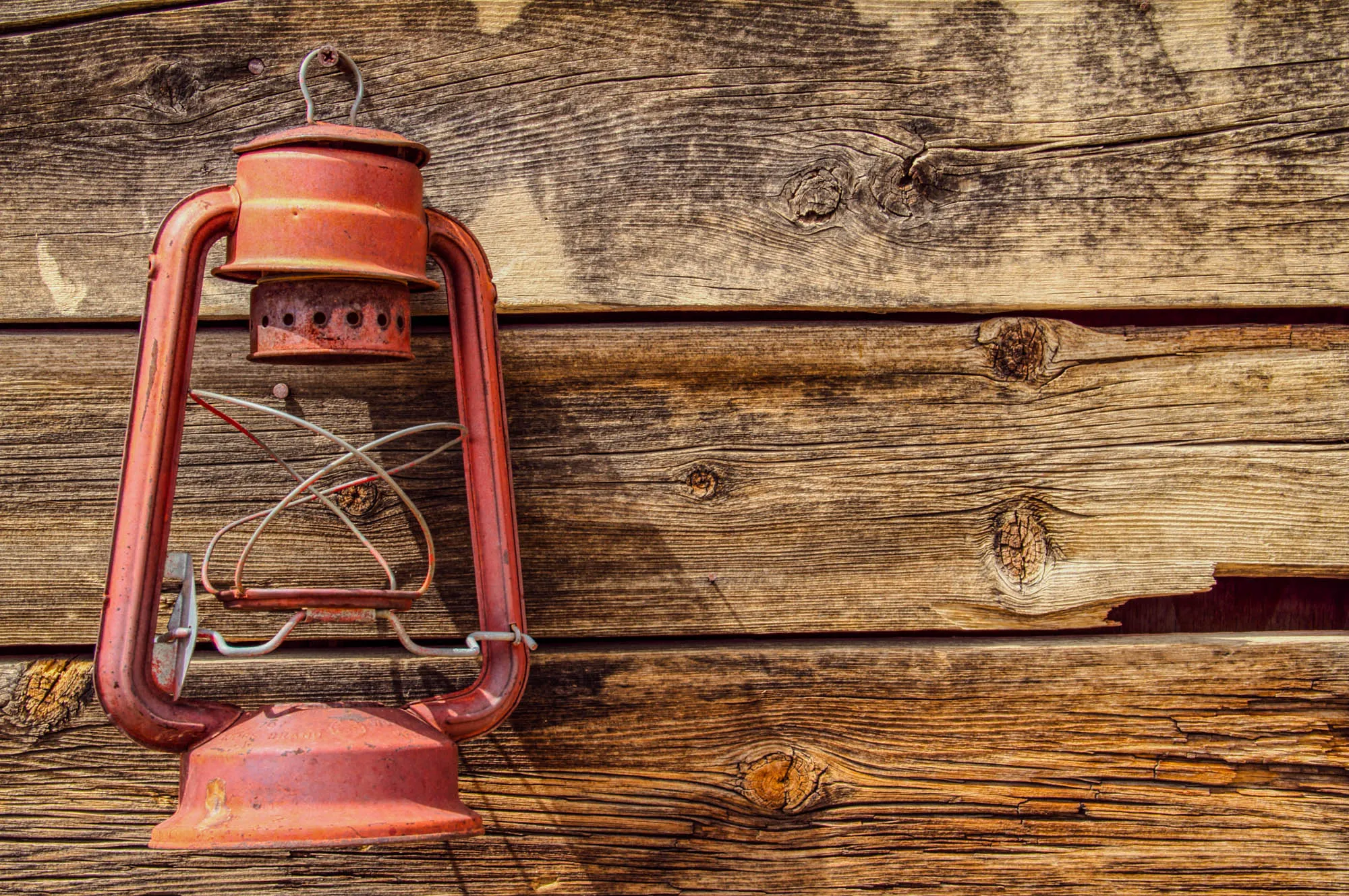 An old rusty red lantern hangs on a weathered wooden wall. The lantern is made of metal with a wire cage around the glass. The wooden wall is made of several horizontal planks of wood. The wood is a light brown color with a lot of knots and cracks. The lantern is on the left side of the image and the wood is on the right side of the image. The lantern is in focus and the wood is out of focus.