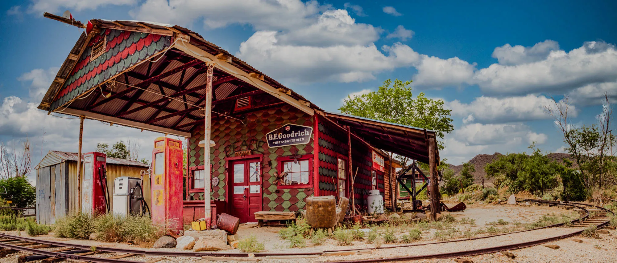 The image shows a weathered, red and green building with a tin roof, a large awning, and a sign that says “B.F. Goodrich Tires-Batteries.”  There are several old gas pumps in front of the building, two of which are red and one that is white. The building is set against a backdrop of hills and trees, with a bright blue sky with fluffy white clouds above. A set of rusty railroad tracks runs in front of the building, curving away to the right.