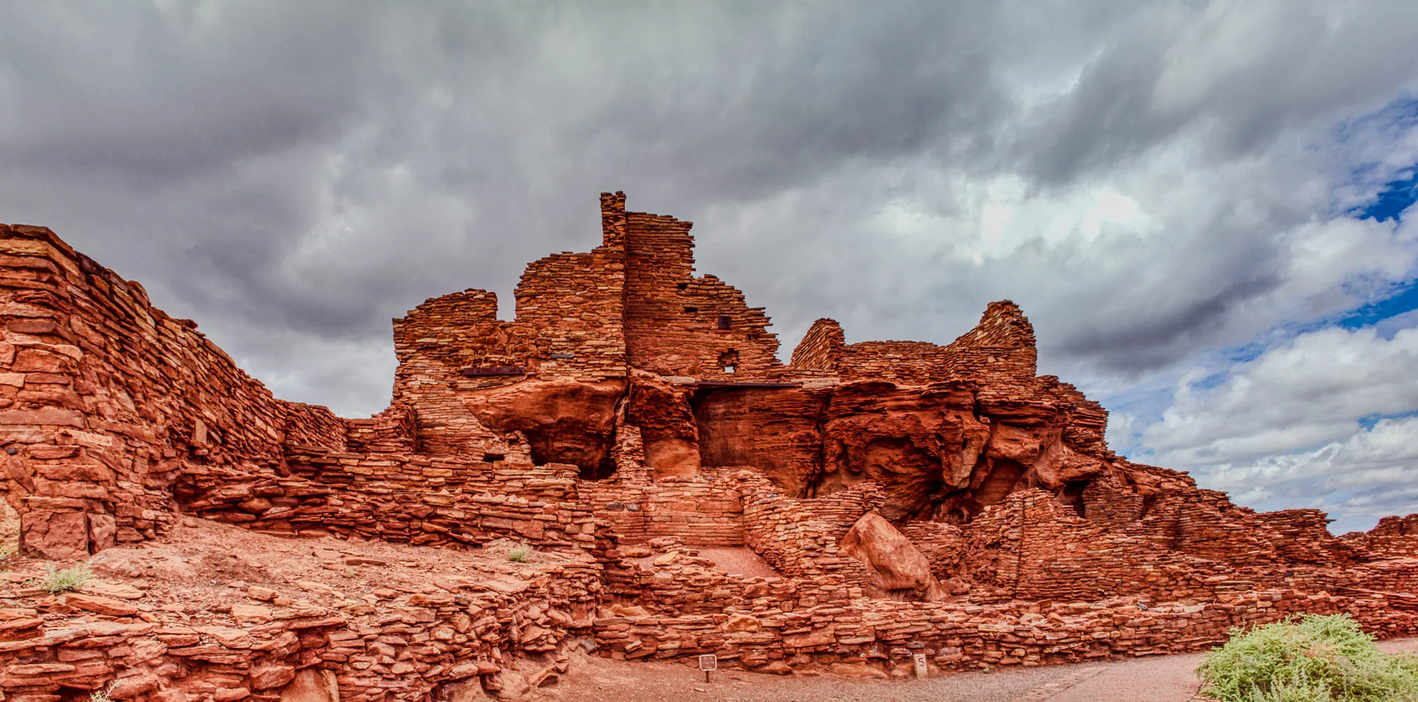 The image shows a large, red-rock ruin, likely a cliff dwelling, set against a partly cloudy sky. The ruins are built into the side of a sandstone cliff, and the rock has eroded in a way that makes it look like a cave, with a large, overhang in the middle. The ruins themselves are constructed of stacked, red-brown rocks. There is a small plant in the lower right corner of the image.  