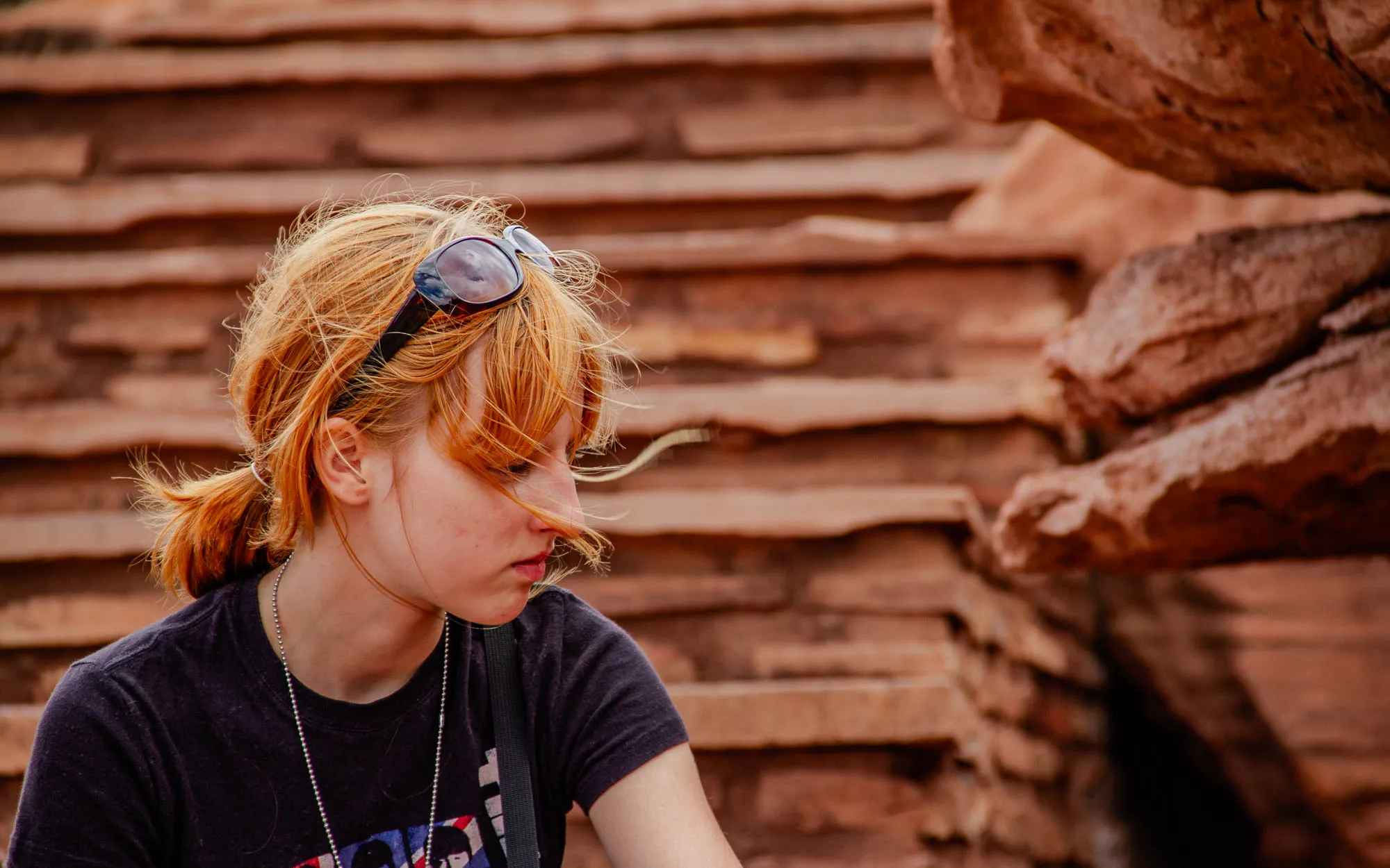 The image shows a young woman with red hair. She is wearing a black t-shirt with a silver chain around her neck. She is wearing dark sunglasses that are pushed up on her head. Her hair is pulled back in a ponytail, and she has a few loose strands falling on her forehead. She is standing in front of a red rock formation, with a row of stairs behind her. She is looking down, and her expression is pensive. The image is likely taken outdoors, and the background is blurred to focus on the woman.