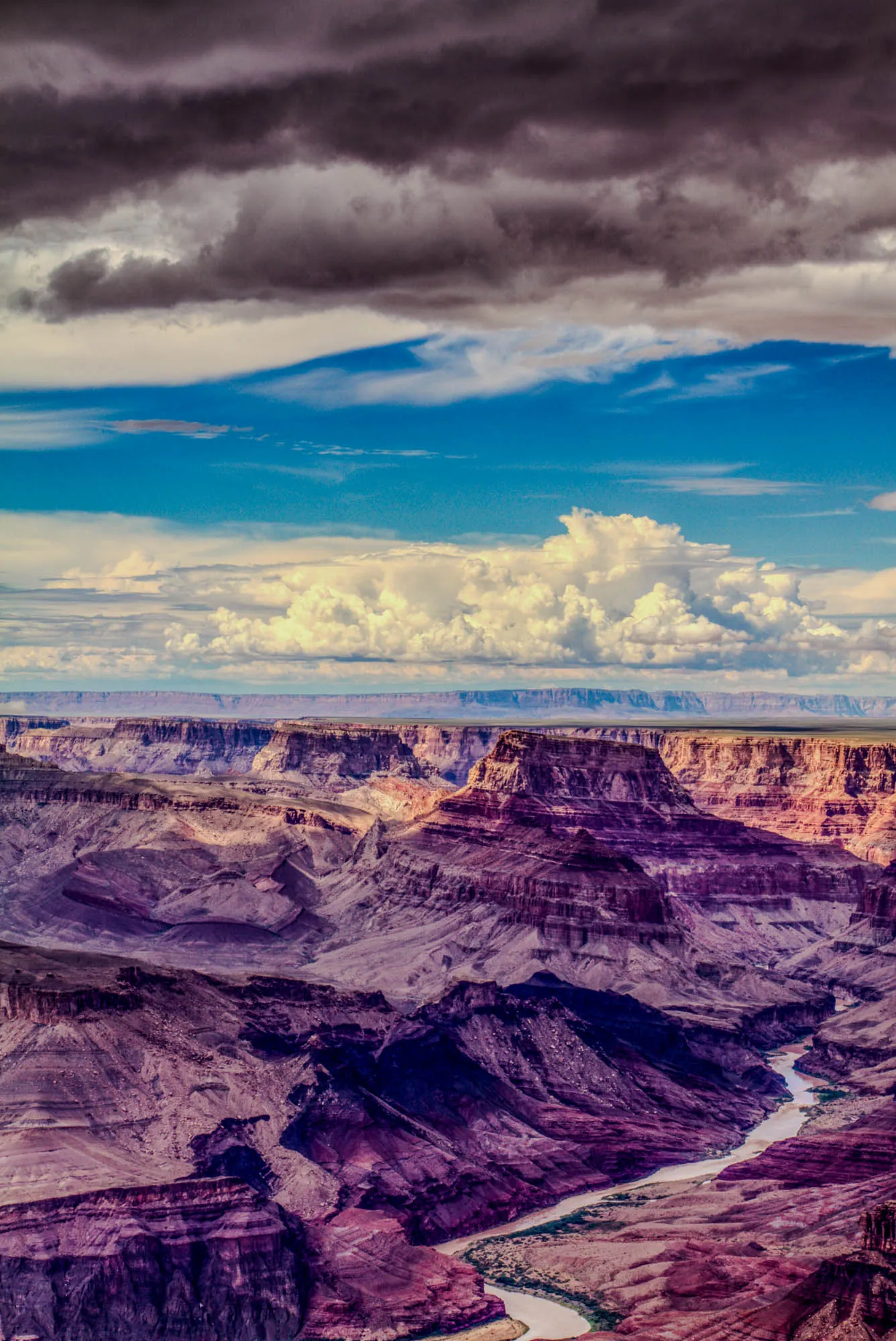 The image shows a breathtaking view of a vast canyon, possibly the Grand Canyon. The sky is a vibrant blue, filled with fluffy white clouds, some of which are darker and more layered, casting a slight shadow over the landscape below. A large, white, puffy cloud dominates the middle of the image, adding depth and dimension to the sky. The canyon itself is a mesmerizing tapestry of brown, red, and purple hues, with layers of rock and sediment exposed, creating a dramatic and textured scene. A silver-colored river winds its way through the canyon floor, adding a sense of movement and life to the composition. The overall impression is one of awe-inspiring grandeur and natural beauty.  
