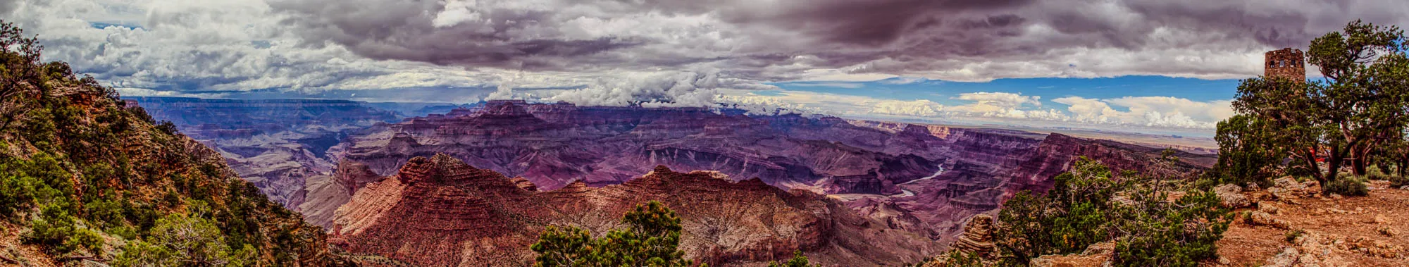 A panoramic view of the Grand Canyon on a cloudy day. In the foreground is a rocky ridge with a few trees growing on it, and the edge of the canyon is visible in the background. The canyon is deep and wide, with many layers of rock visible. In the distance, a small stone tower can be seen, partially obscured by the trees.  The sky is mostly overcast, with large, puffy clouds, but there is a small patch of blue sky visible near the horizon. The image is taken from a high vantage point. The overall tone of the image is dark and moody.