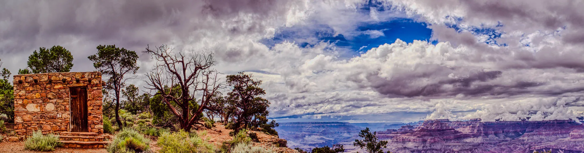 A stone building with a wooden door stands on a cliff overlooking a vast canyon. The building is made of rough-hewn stones and has a low, pitched roof. The door is dark brown and has a simple design. The building is surrounded by trees and shrubs. In the distance, the canyon stretches out in a maze of colorful rock formations. The sky is a mix of dark gray clouds and bright blue patches. The clouds are thick and heavy, casting shadows across the canyon floor. The image is a stunning display of the natural beauty of the Grand Canyon.