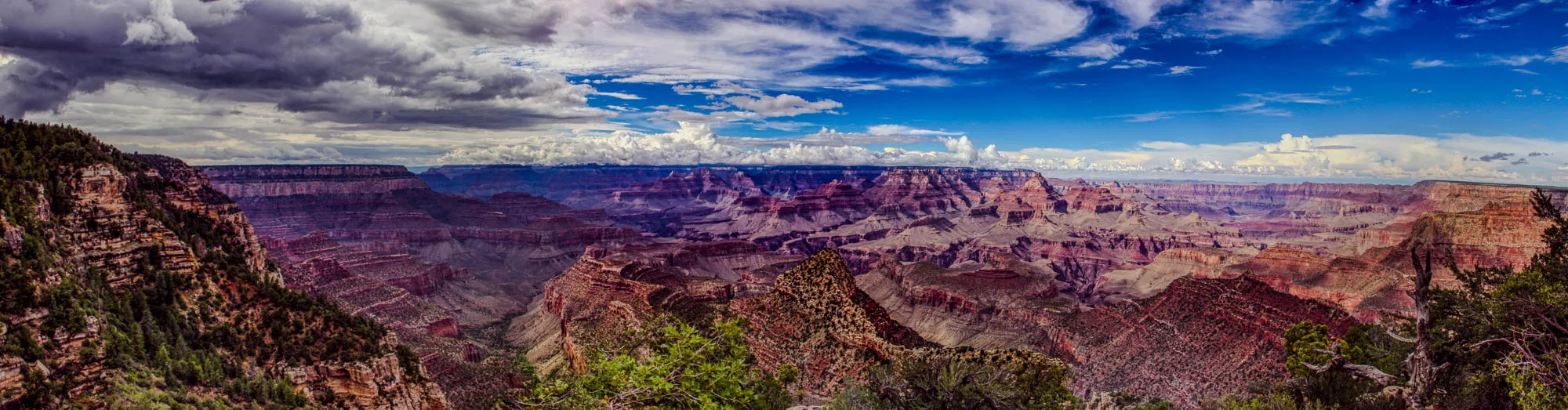 A panoramic view of the Grand Canyon on a partly cloudy day. The viewer is looking out from a cliff at the edge of the canyon, with a view to the south, looking down into the canyon. The canyon is vast and deep, with red rock cliffs that are layered and textured. In the foreground, there are trees growing along the edge of the cliff. The sky is blue with fluffy white clouds. The image is a beautiful example of the natural beauty of the Grand Canyon.