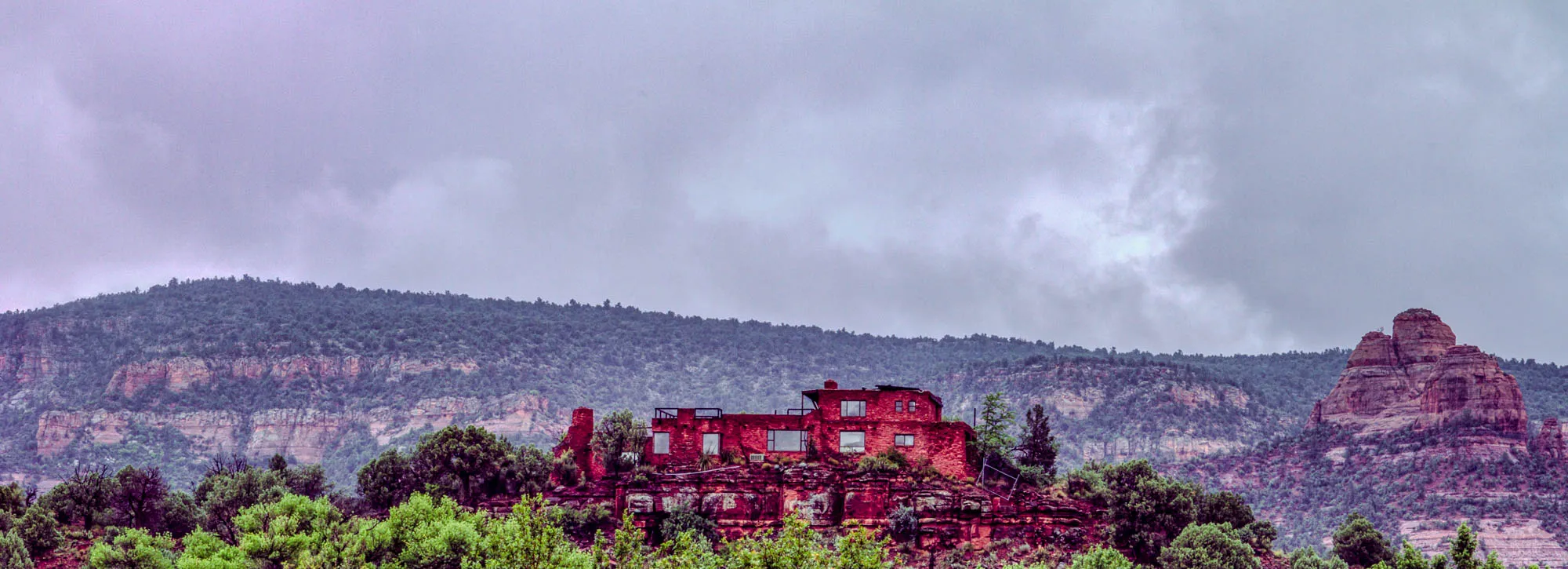 The image shows a red brick building nestled on a cliffside. The building is a few stories tall, with a flat roof and several windows. In front of the building are several green trees and bushes, and the ground is a mix of red rock and dirt.  In the distance, there are large rocky hills with a lot of green trees and bushes. There is a gray overcast sky with wispy clouds. The overall feeling of the image is one of peace and tranquility. 
