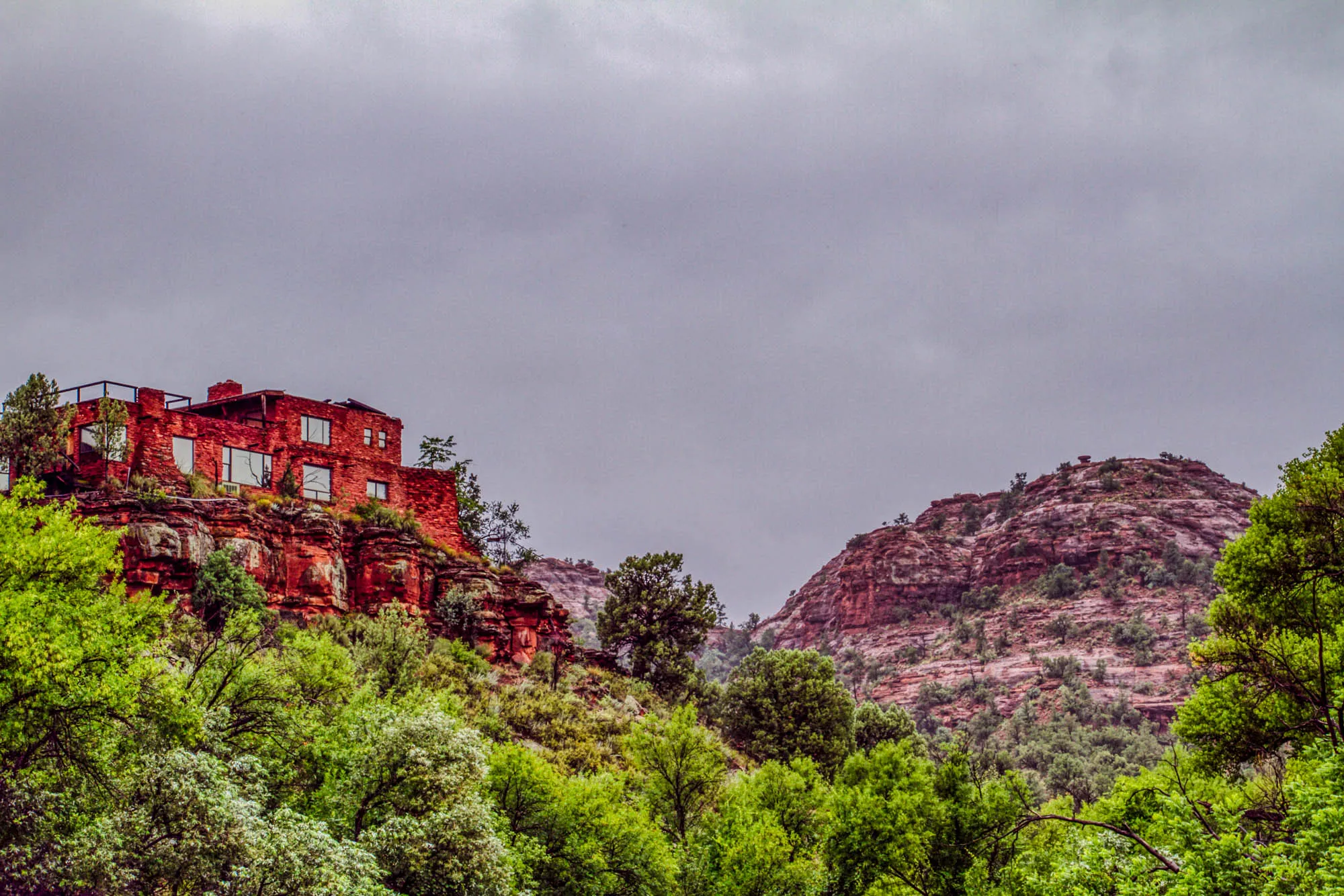 The image depicts a red brick house perched on the edge of a steep cliff. The house has multiple windows, a flat roof, and is surrounded by lush green trees. The cliff is composed of reddish-brown rock with patches of green vegetation. In the background, a second, larger cliff with a similar reddish-brown coloring rises up against a hazy, gray sky. The overall atmosphere is one of dramatic beauty and a sense of isolation.