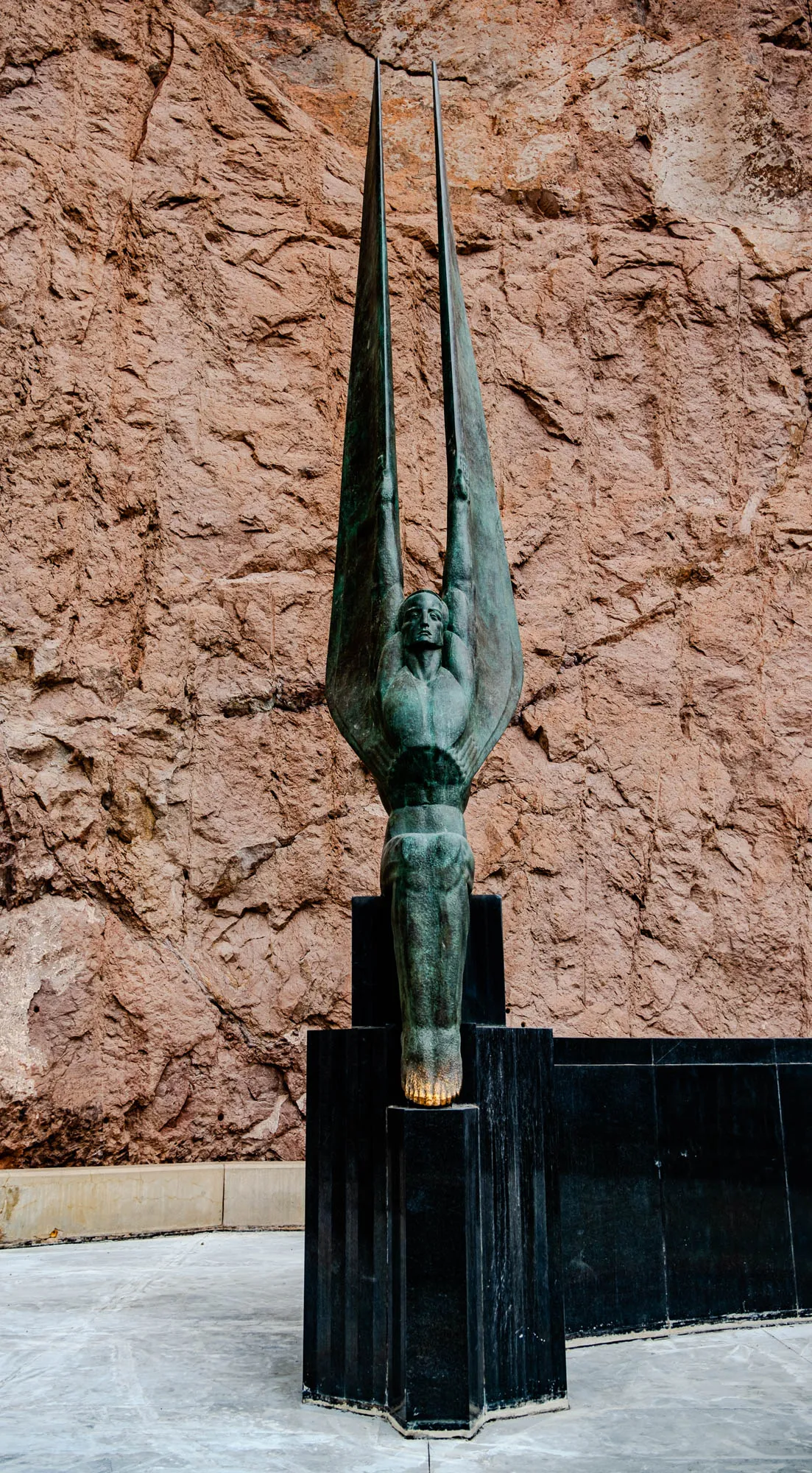 The image depicts a bronze sculpture of a man with outstretched arms, resembling wings, against a backdrop of a rough, textured reddish-brown rock face. The sculpture stands on a black rectangular pedestal, with a lighter-colored stone ledge in front of it. The sculpture is weathered and has a green patina, with a slight sheen. The man's face is visible, with a determined expression. The statue is positioned on a large flat concrete surface, with a slight perspective slant in the foreground. The background rock face is a natural rock formation, with many cracks and crevices, creating a sense of age and grandeur.  The statue seems to symbolize a sense of aspiration or soaring towards the sky.  It is likely a public monument or memorial, reflecting the grand scale of the surrounding landscape.  The sculpture stands in contrast to the natural ruggedness of the rock face, creating a juxtaposition of human creation against the forces of nature.