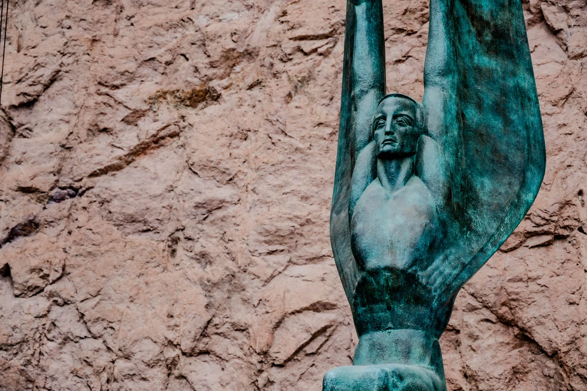 A bronze statue of a man with wings raised above his head, the man is looking straight ahead. He is standing in front of a rough, textured brown rock wall. The sculpture is a teal-green color, and the man's face has a serious expression.  The man's wings are flowing upward.  The statue is sculpted in an art deco style.