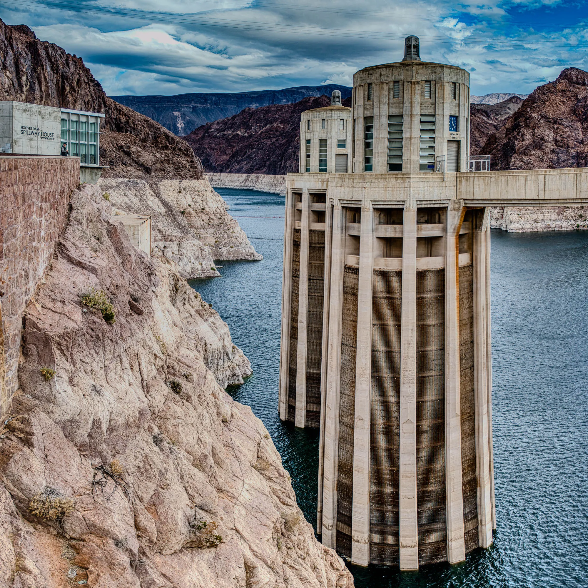 The image shows a view of the Hoover Dam spillway towers from the dam. The two towers are made of concrete and are positioned next to each other. The towers are tall and have a cylindrical shape, with a series of horizontal lines running across their surface. The towers are partially submerged in blue water. 

To the left of the towers, a cliff face is visible, made of tan-colored rock. The rock face is uneven, with crevices and outcroppings. There is a small, rectangular building at the top of the cliff, with a sign that reads "Hoover Dam Spillway House, Event Center". The background is made up of rocky mountains and a blue sky with white clouds.