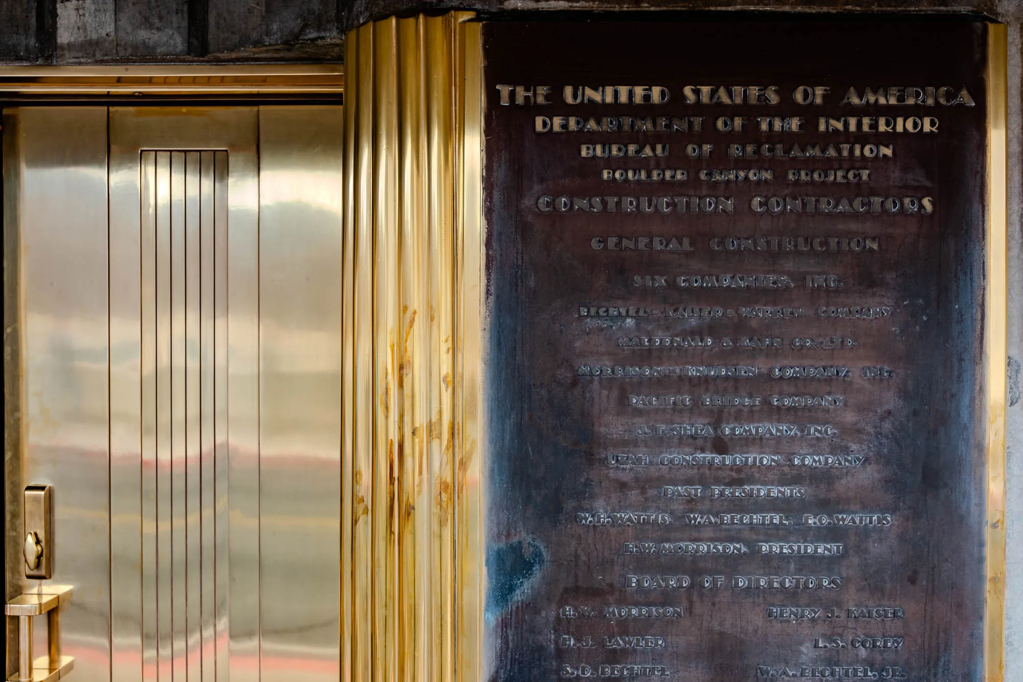 The image is of a brass door with a plaque on the right. The door is shiny and has vertical grooves running the length of it. The door has a handle and lock on the left side of the door. The plaque is dark gray and has gold text. The text on the plaque is: “The United States of America Department of the Interior Bureau of Reclamation Boulder Canyon Project Construction Contractors General Construction Six Companies, Inc. Bechtel-Kaiser-Warren Company MacDonald and Kahn Co., Ltd. Morrison-Knudsen Company, Inc. Pacific Bridge Company J. F. Shea Company, Inc. Utah Construction Company Past Presidents W.H. Wattis, W.A. Bechtel, E.O. Wattis H.W. Morrison, President Board of Directors H.W. Morrison Henry J. Kaiser H. J. Lawler L.S. Corey S.D. Bechtel W.A. Bechtel, Jr.”