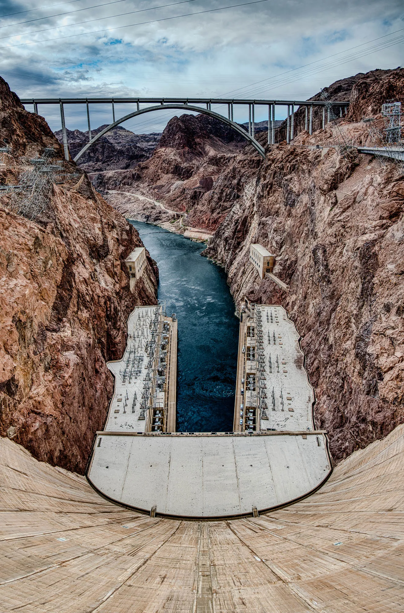 The image shows a view of Hoover Dam from above.  The dam is made of concrete and has a curved shape. There is a river running through the middle of the image, and the dam is built between two rocky cliffs. In the distance, there is a bridge that goes over the river and the cliffs. The river is blue and the water is choppy. The sky is grey with white clouds. The cliff faces are red brown and the rocks have lots of texture.  The top of the dam has a large flat space with a few white cars on it. The dam appears to be quite tall. There is an arch bridge crossing the canyon a ways off. It has support beams in the middle.  There is a light brown colored bridge across the top of the dam with a walkway, this bridge is not arched.  There are two buildings on the sides of the dam and many cables with metal posts on the sides of the dam. 
