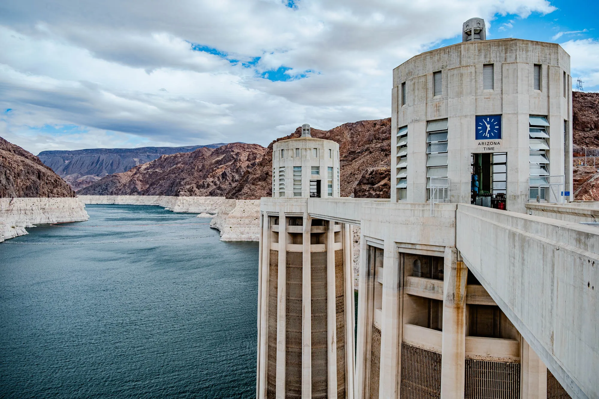 The image shows a view from the top of Hoover Dam. The dam is made of concrete and has a large, rectangular opening in the middle of the dam. There are two cylindrical towers on either side of the opening. The tower to the right of the image has a clock face that reads "Arizona Time." The towers are connected by a walkway. The water is a deep blue and the dam is surrounded by rocky mountains. The sky is a mix of blue and white clouds.