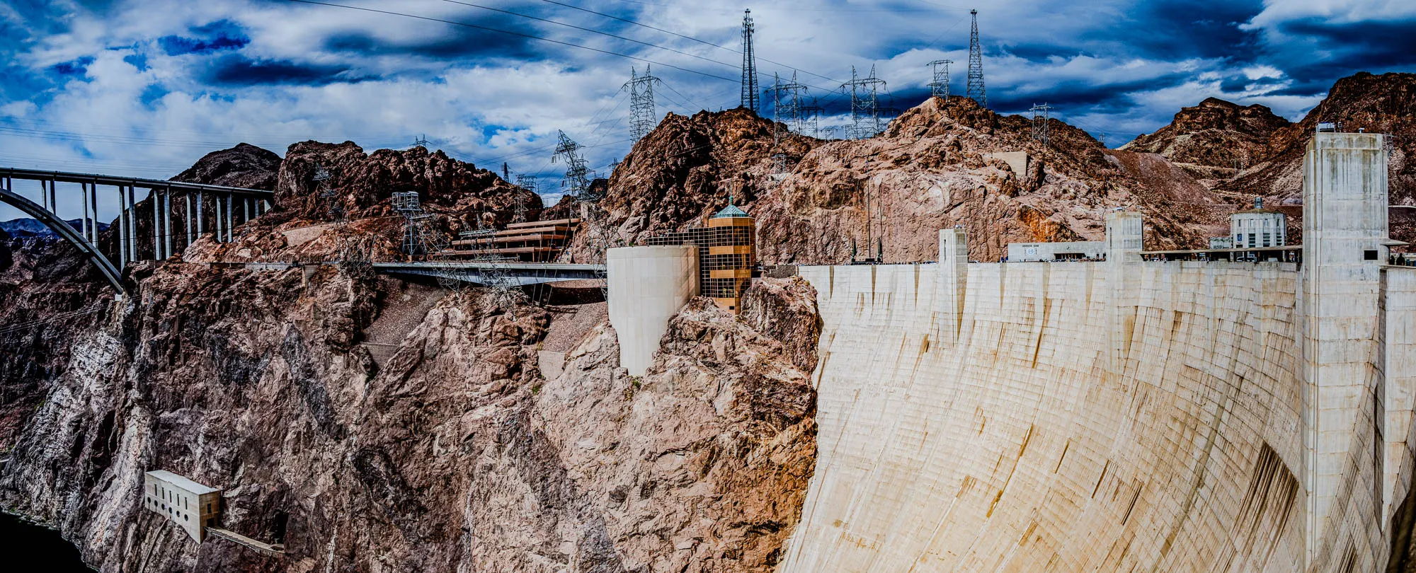 A view from a high vantage point looking down at the Hoover Dam. The dam is made of concrete and has a curved shape. In the foreground, a portion of the dam is visible, and behind it is a large, rocky, reddish-brown mountain. The top of the mountain is dotted with power lines and towers. In the distance, there is a bridge, and the sky is cloudy and blue.