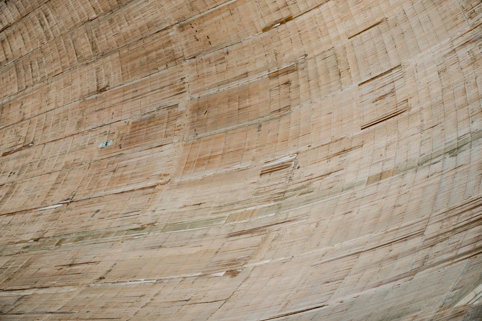 The image is a close-up of the inside of a concrete dam. The concrete is a light brown color and has a textured, rough surface. The dam curves inward and is made up of horizontal layers of concrete, giving the appearance of a  smooth, gentle slope. The dam is likely very large and imposing, and the image captures a small section of its interior.  There are no other details in the image.