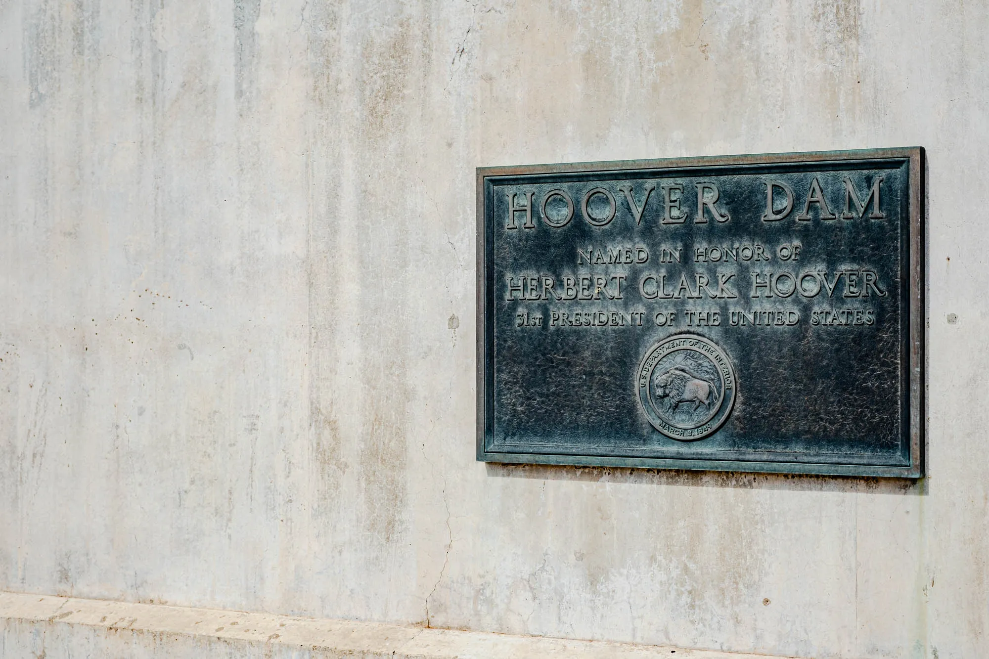 The image shows a plaque mounted on a concrete wall. The plaque is a dark bronze color with a raised silver border. It reads "HOOVER DAM" in large letters at the top, followed by "NAMED IN HONOR OF" in a smaller font. The next line reads "HERBERT CLARK HOOVER" in a larger font, and the final line reads "31st PRESIDENT OF THE UNITED STATES" in the smaller font. Below the text is a seal with the words "U.S. DEPARTMENT OF THE INTERIOR" and "MARCH 3, 1849" inscribed within it. The seal depicts a bison. The background of the image is the plain concrete wall.