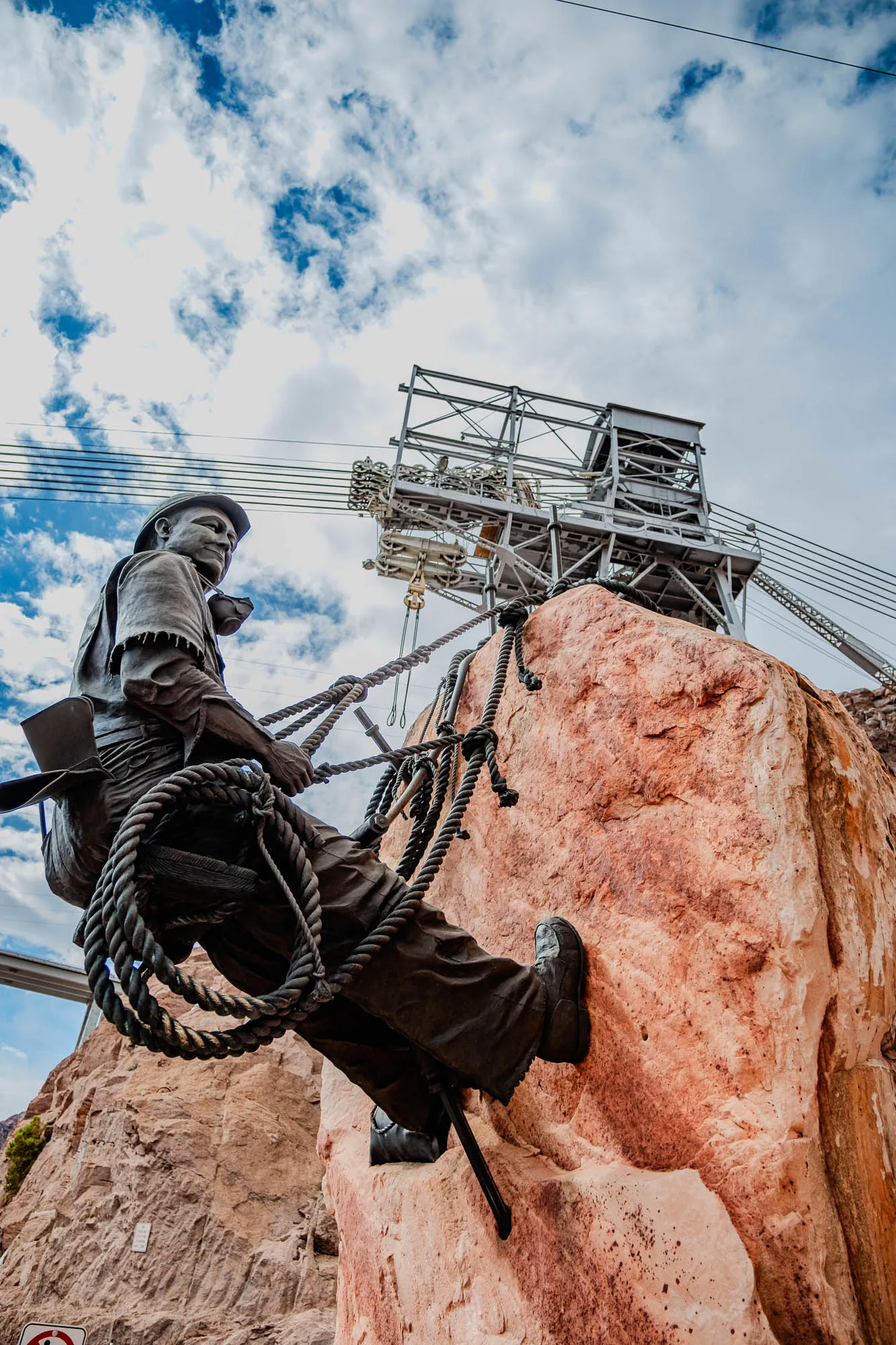 The image shows a bronze statue of a man climbing a rock face. He is wearing a hard hat, work boots, and a jacket. He is secured to the rock with ropes. The rock is a light orange color and is textured.  The man is looking up at a metal structure that is out of frame at the top of the image.  The background is a blue sky with white clouds. There is a white metal structure in the background with wires and ropes attached.  The statue is set against a backdrop of a rocky canyon wall.  A sign is visible at the bottom of the image.  The statue appears to be a memorial to the workers who built the nearby dam.