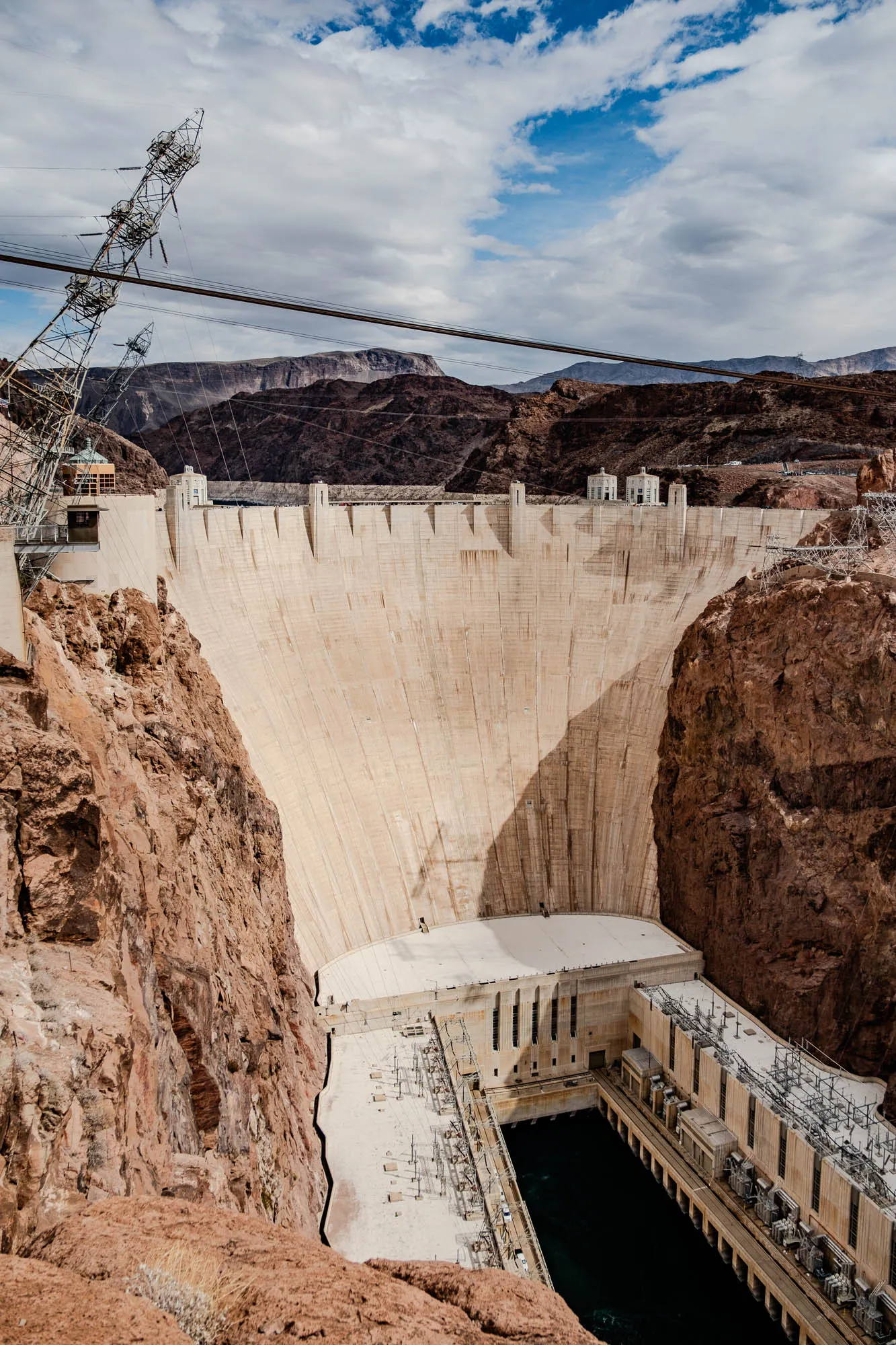 The image shows a large concrete dam with a curved face. The dam is set in a rocky canyon, with the right side of the image showing a sheer cliff of brown rock. The dam is very large and extends across most of the image. The dam is made of concrete blocks and has a wide flat top with a large pool of water at the base. There are numerous power lines running across the top of the dam and above it. The sky is blue with white clouds.