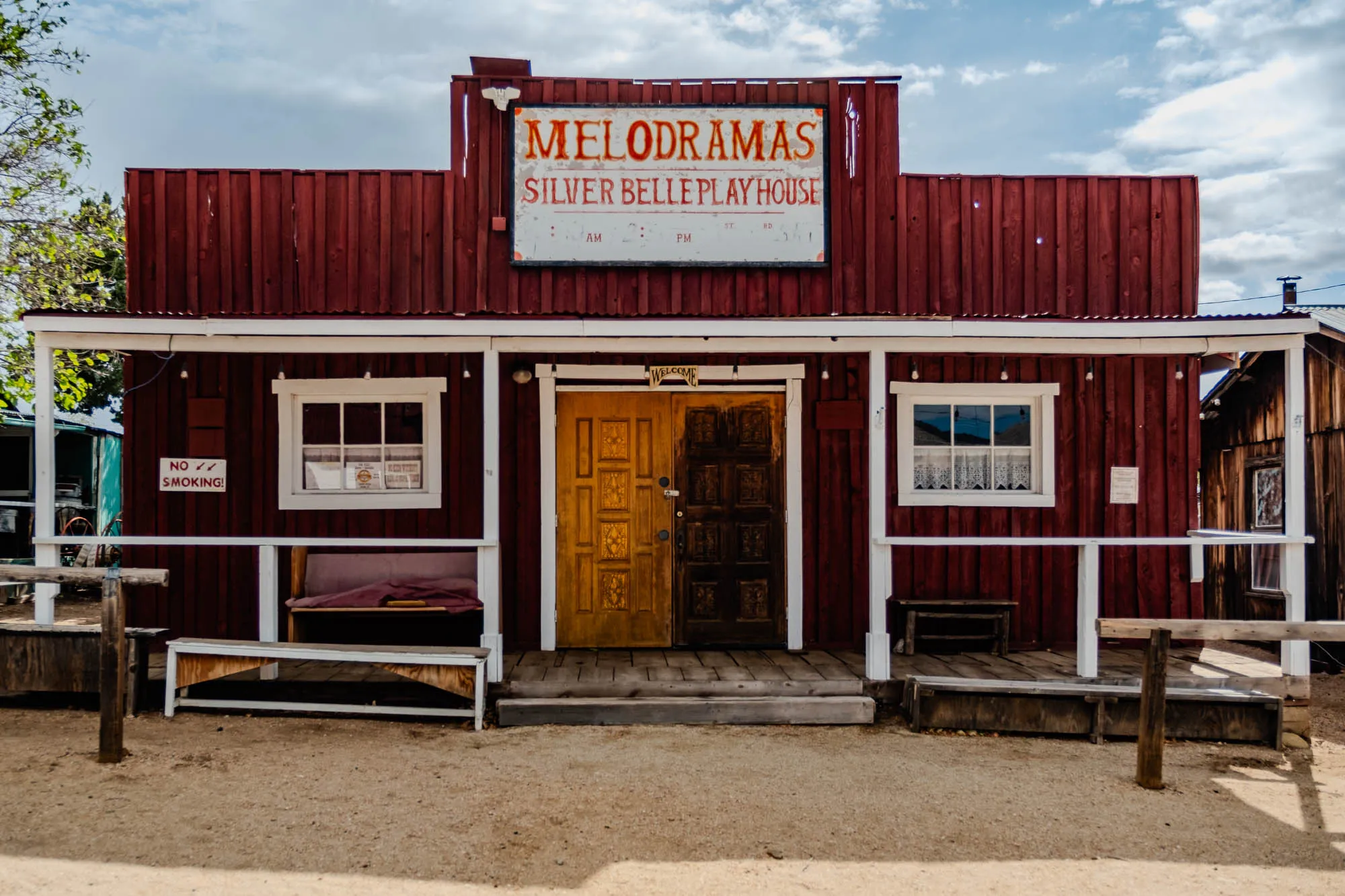 The image shows a red wooden building with a white porch and a sign above the door. The sign reads "Melodramas Silver Belle Playhouse" and is located in the center of the building. There are two doors leading into the building, one is gold and the other is dark wood.  A window is visible on each side of the doors.  A no smoking sign is on the left side of the image, next to a bench.  A bench is also on the right side of the image, with a  wooden railing. The image is taken on a sunny day, and there are blue skies with white clouds in the background.  The ground is dirt.  The image is taken at a slight angle from the ground.